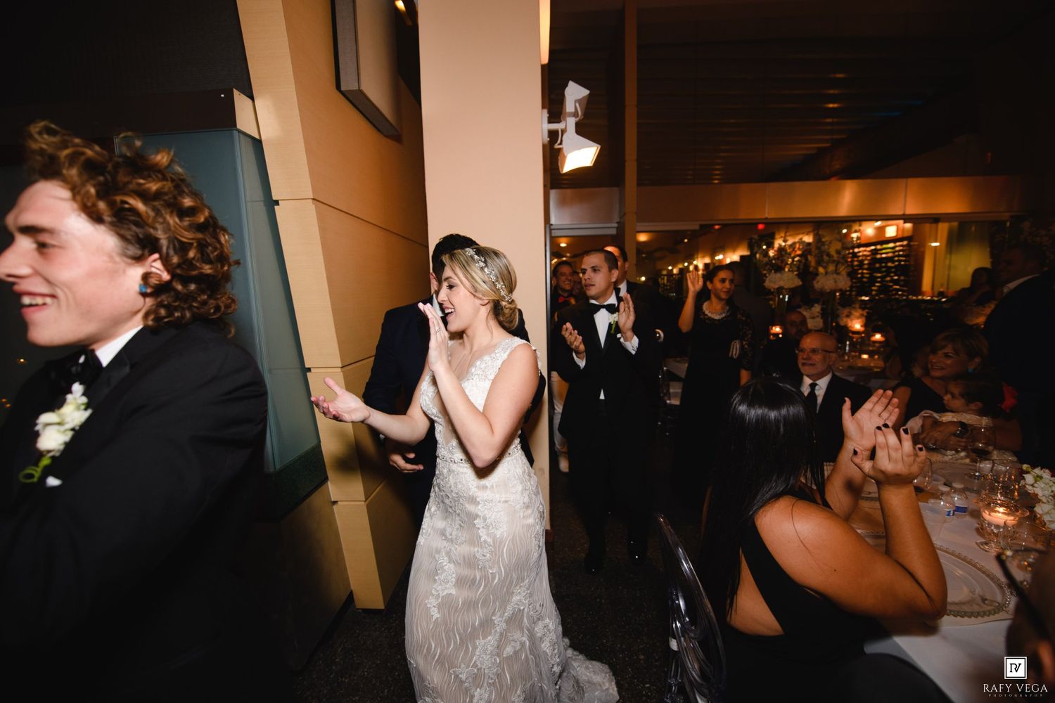 Bride in lace gown laughs joyfully as wedding guests applaud during a candlelit reception entrance.