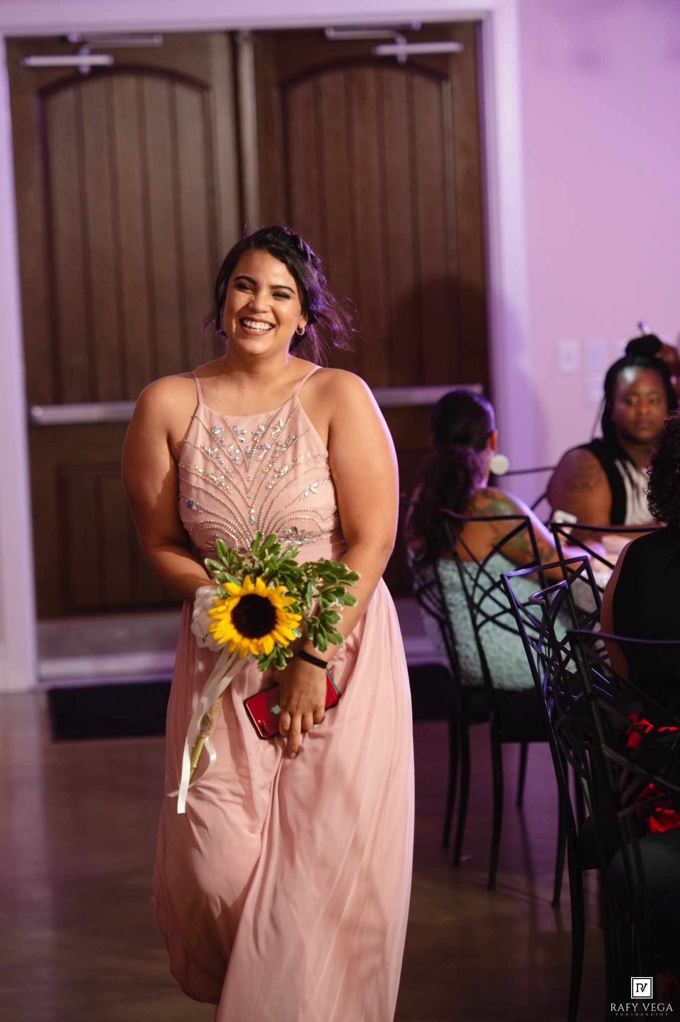 Smiling bridesmaid in pink beaded gown holding sunflower bouquet walks at elegant wedding reception.