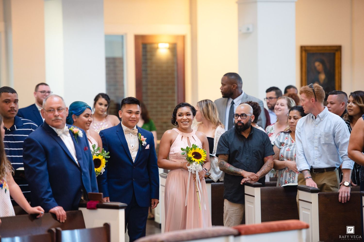 Wedding procession in a church with bride holding sunflowers, groom, and guests in pews during a ceremony.