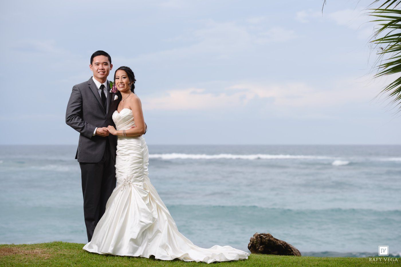Bride and groom posing together on a tropical oceanfront lawn, bride in mermaid gown, groom in gray suit.
