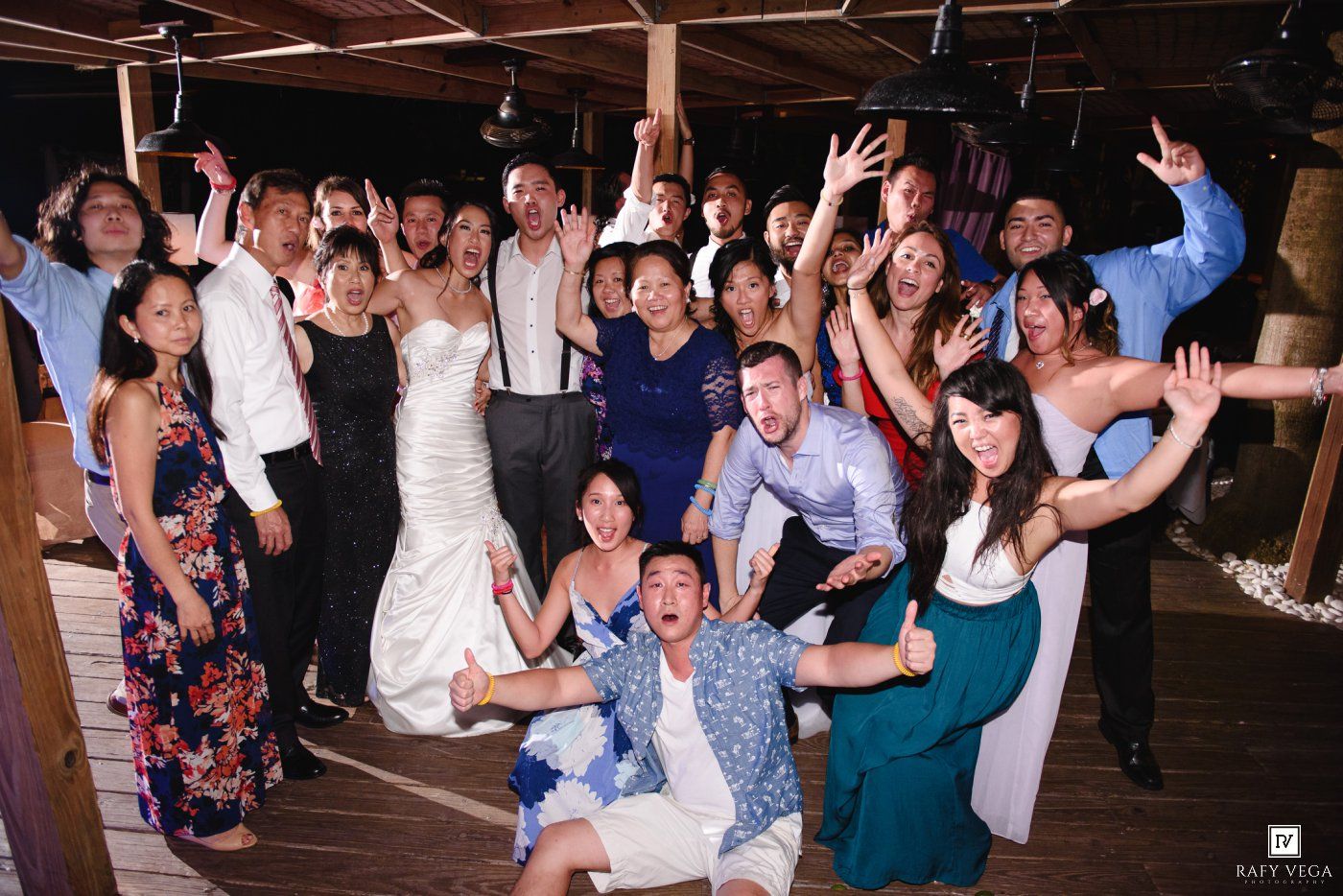 Joyful wedding reception group photo with bride and groom surrounded by celebrating guests on a dance floor.