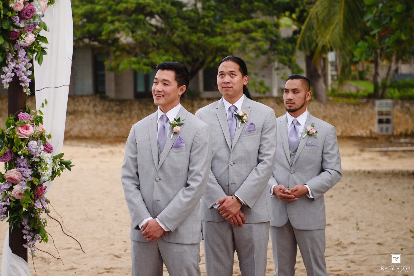 Three groomsmen in light gray suits with purple boutonnieres stand at a beach wedding ceremony altar.