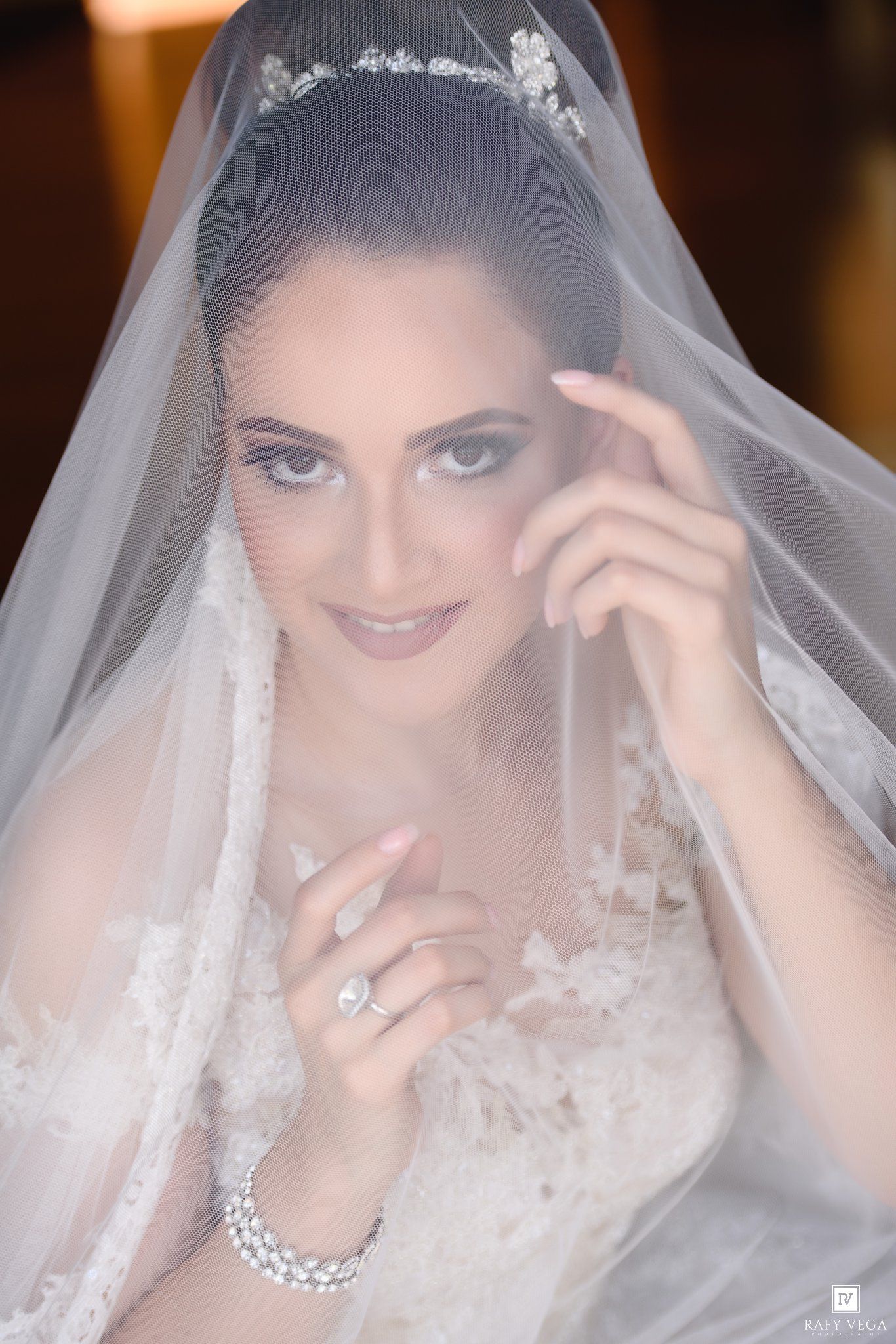 Elegant bride in lace gown and floral tiara, draped in sheer veil, showcasing diamond ring and bracelet.