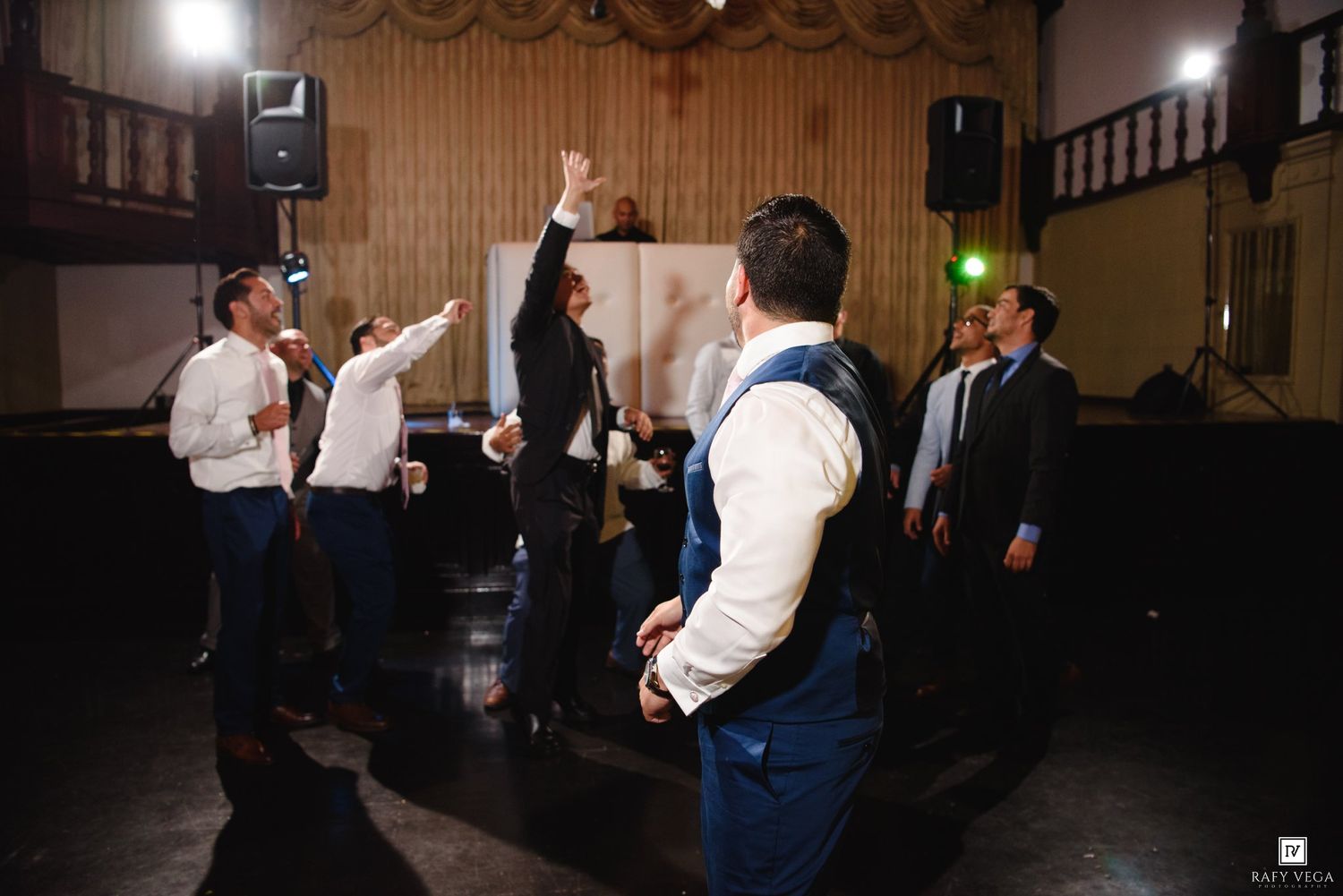 Men in formal attire dancing and celebrating at a wedding reception in a dimly lit venue.