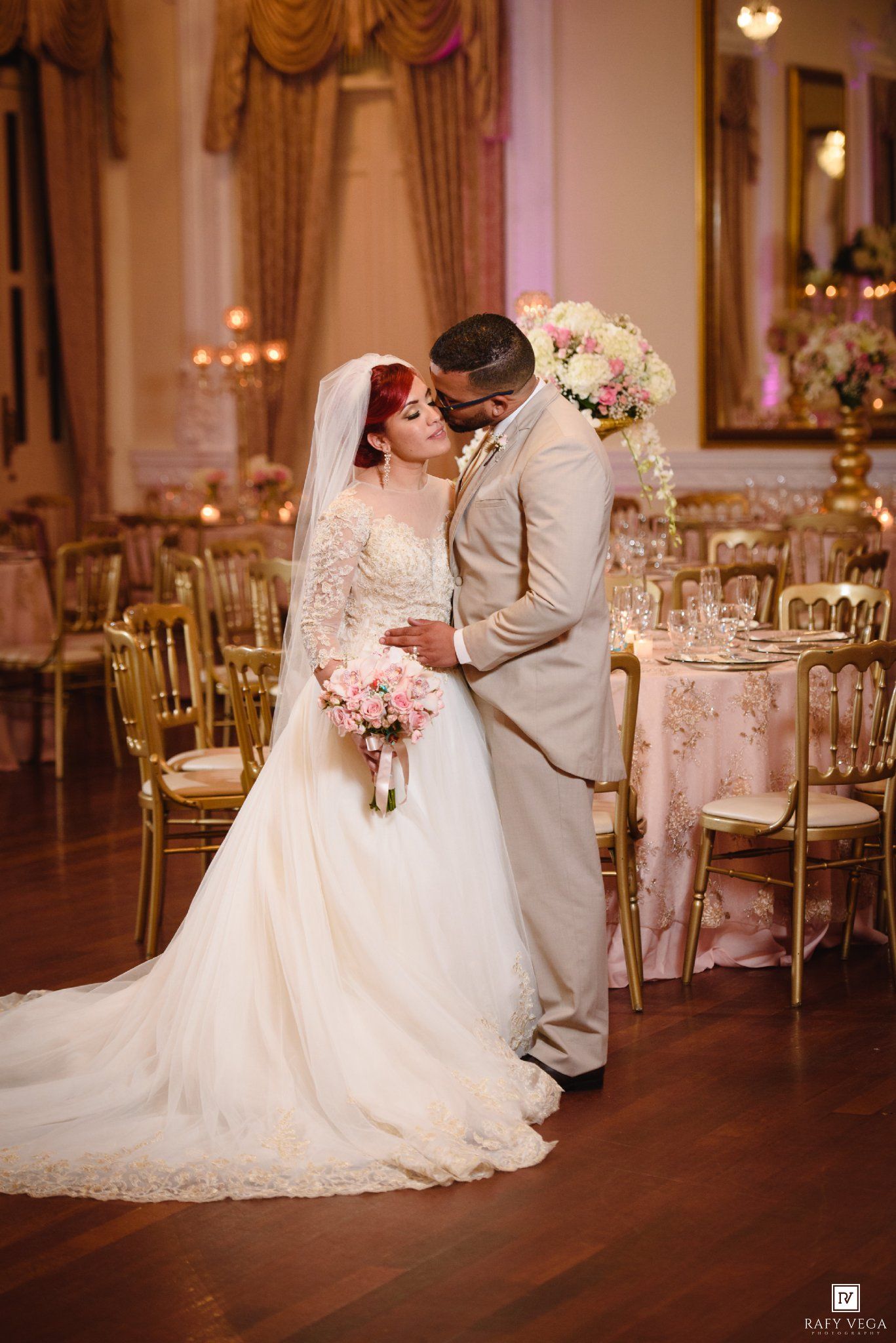 Bride in white lace gown and groom in tan suit share romantic moment in elegant gold ballroom wedding reception.