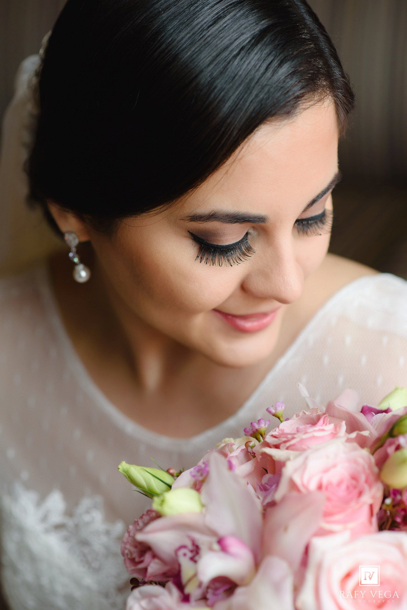 Elegant bride with dark hair holding a pink rose bouquet, wearing pearl earrings and a white lace dress on her wedding day.