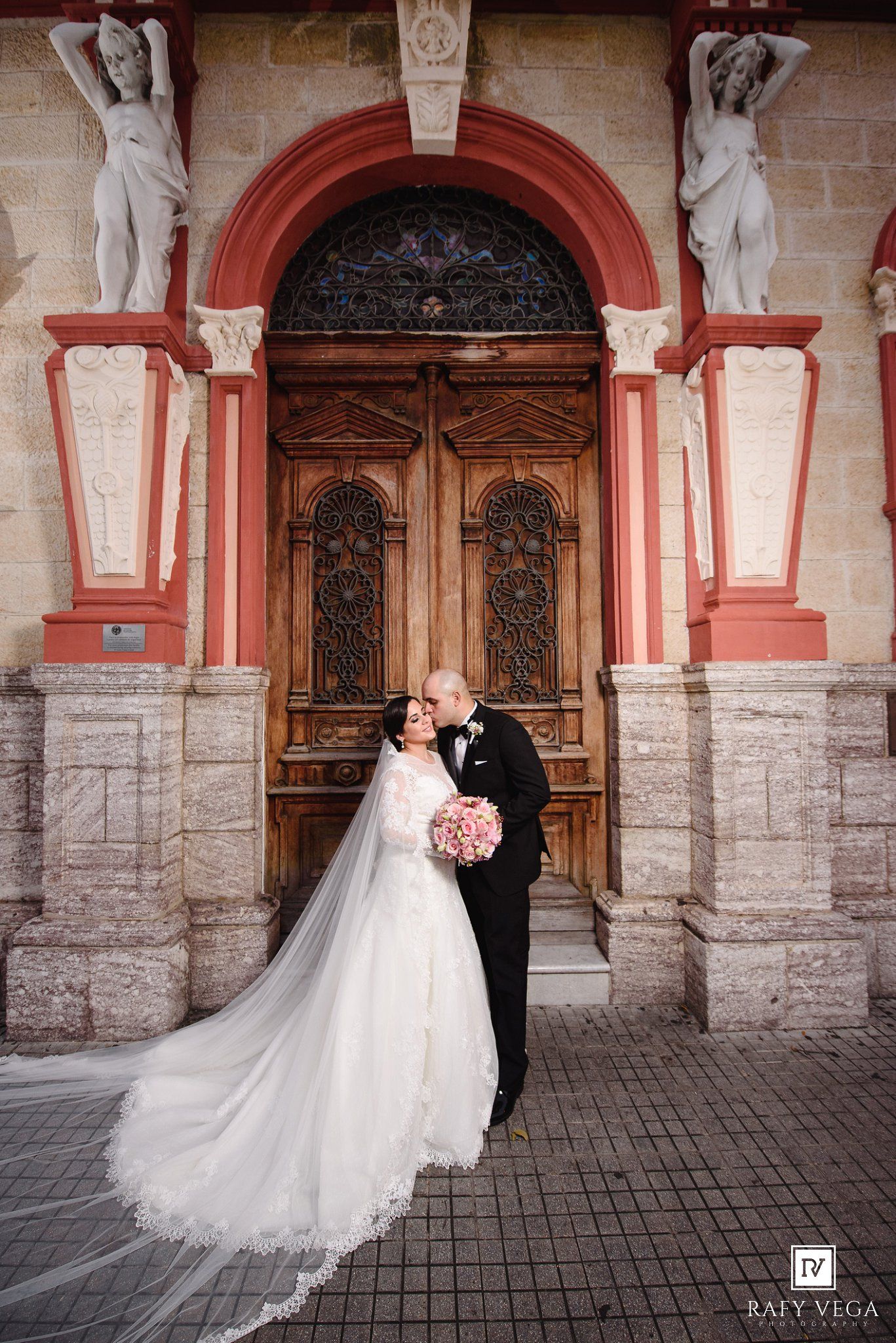 Bride and groom share a romantic kiss in front of an ornate wooden door with classical statues at a wedding venue.