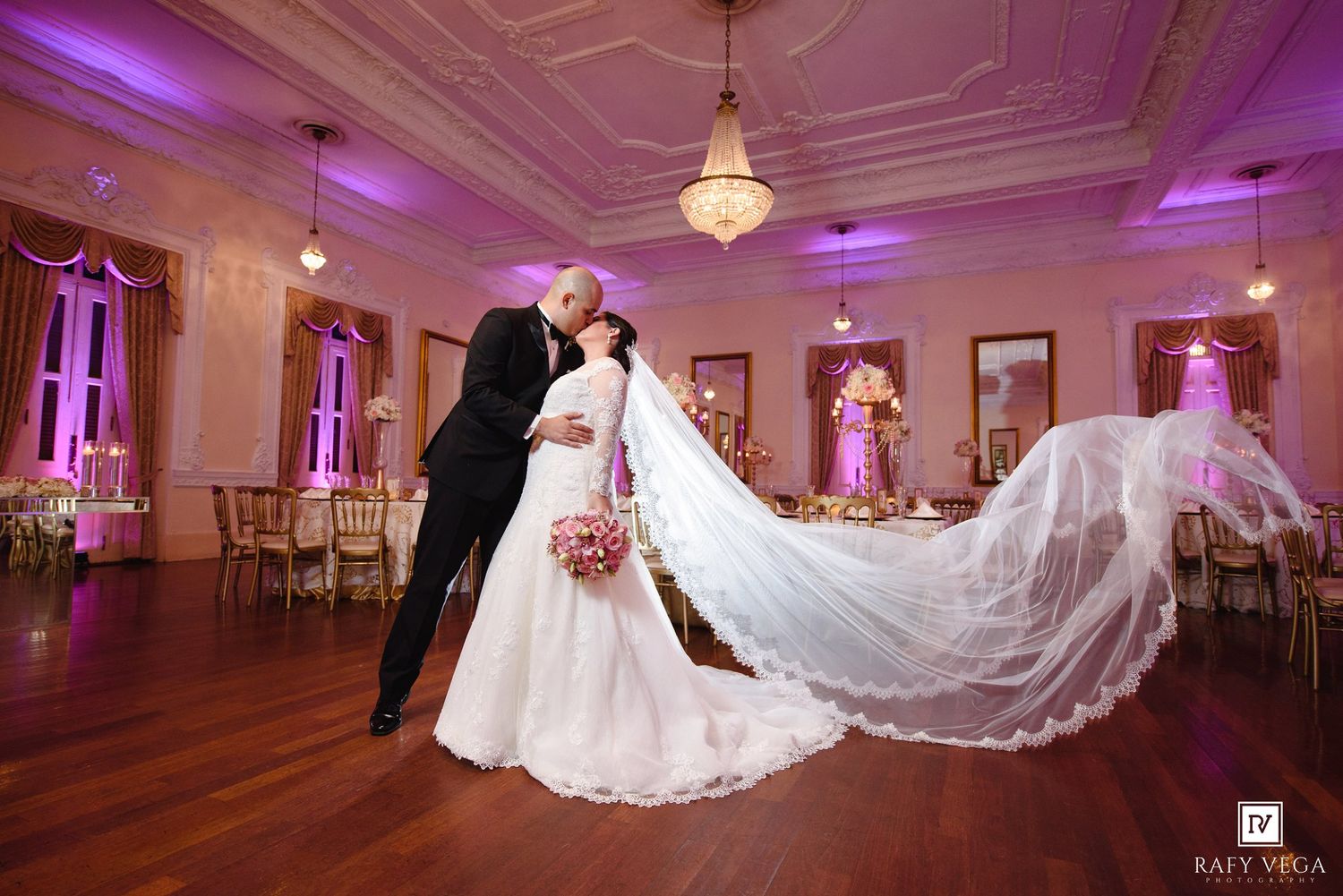 Bride and groom share romantic first dance in elegant ballroom with pink uplighting and flowing cathedral veil.