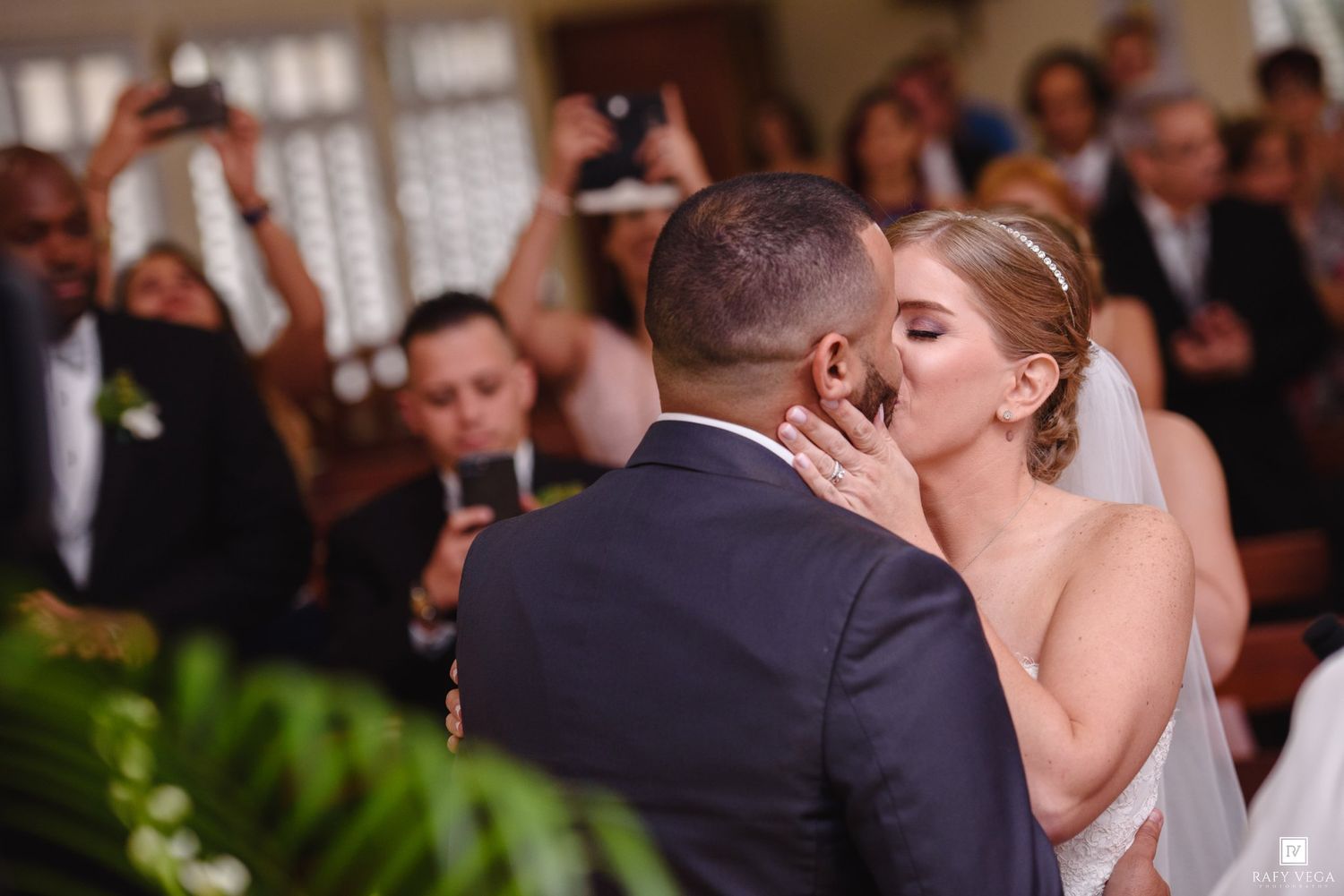 Bride and groom share their first kiss at wedding ceremony while guests watch and photograph the romantic moment.