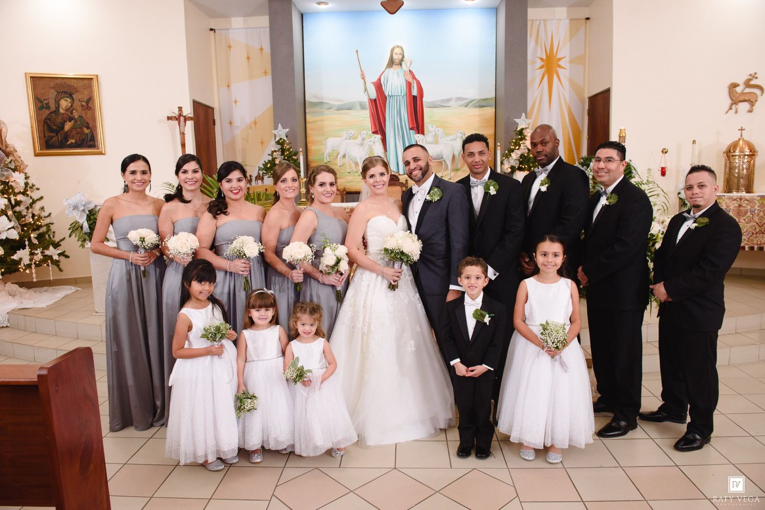 Wedding party poses in a church with bridesmaids in gray dresses, groomsmen, flower girls, and bride and groom at center.