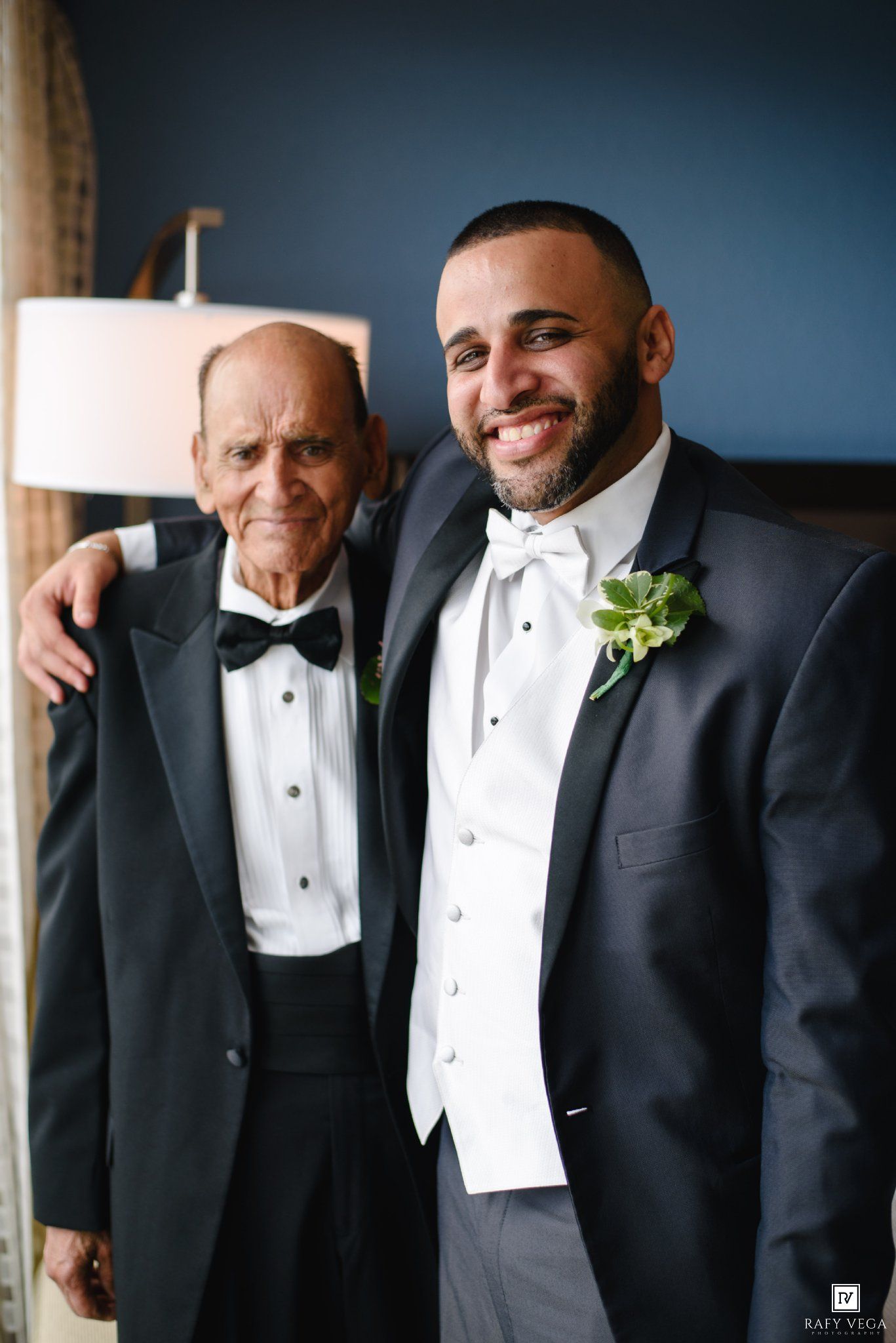 Two men in tuxedos with bow ties pose together at a wedding, one wearing a white vest and floral boutonniere.