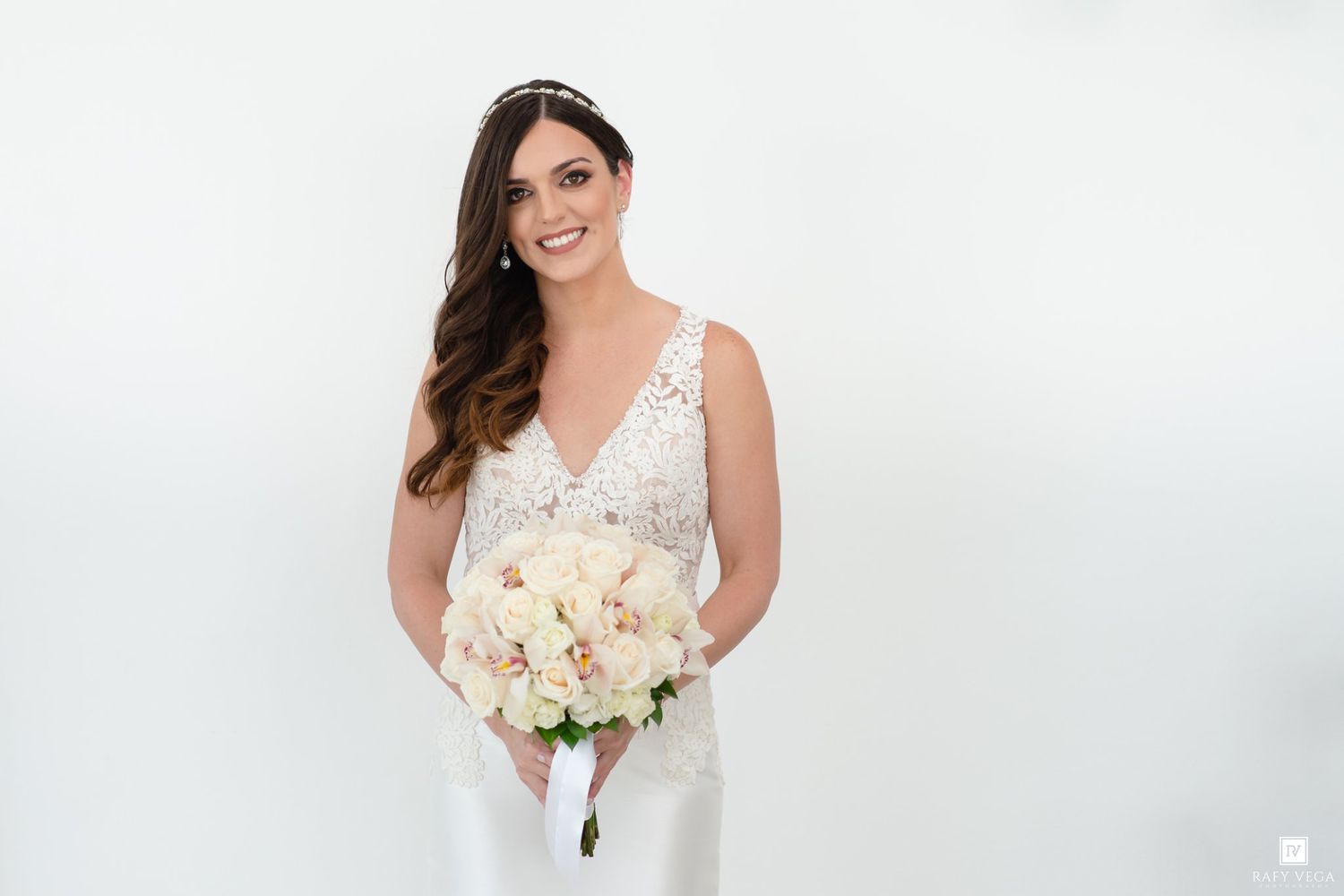 Smiling bride in lace wedding gown holds white rose bouquet against white background.