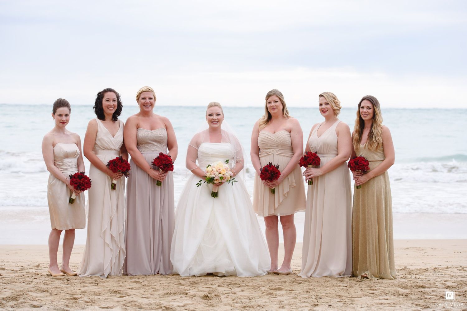 Bride and six bridesmaids in champagne gowns holding red bouquets pose on a sandy beach with ocean waves behind them.