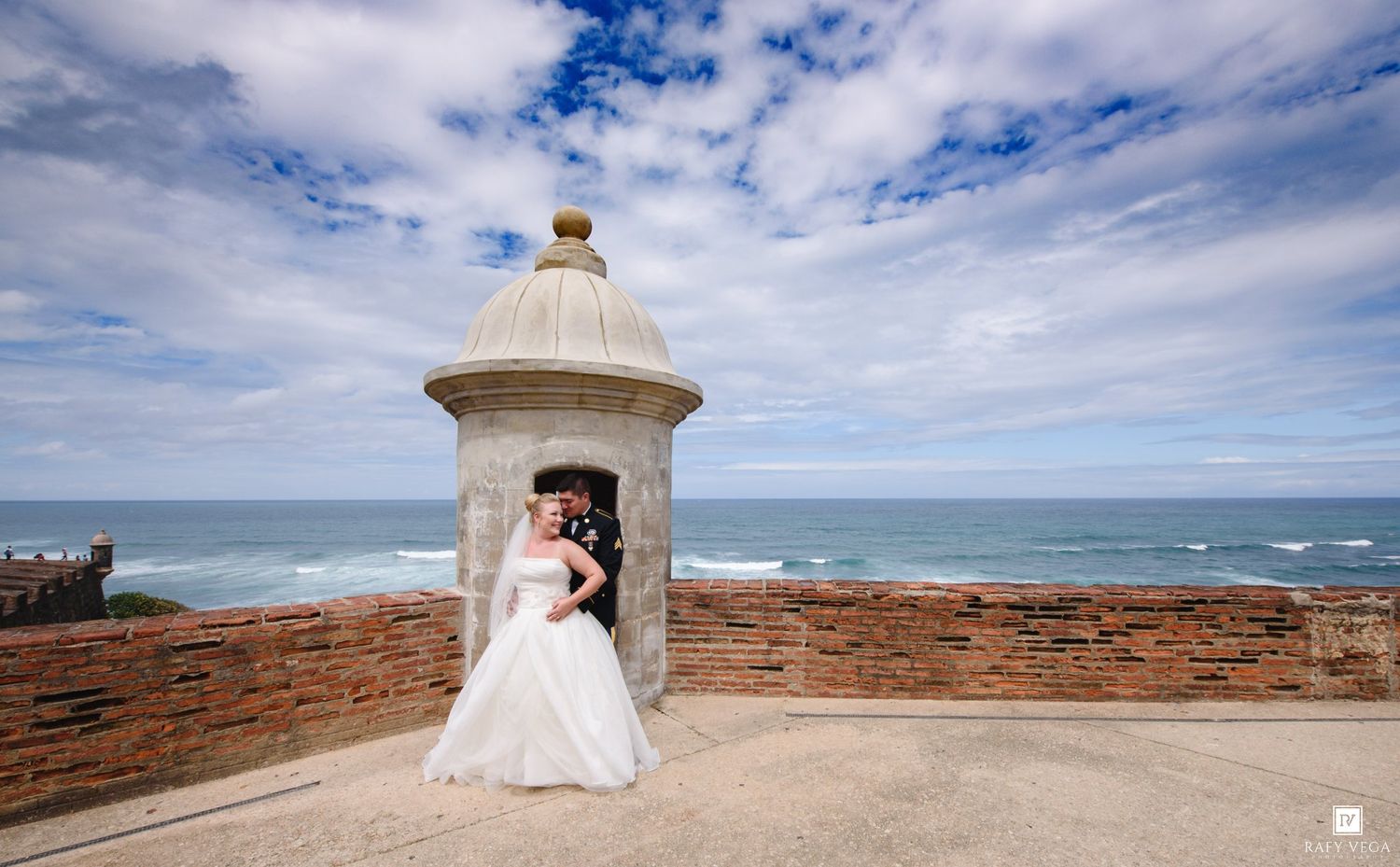 Bride and groom embrace at historic El Morro fort in San Juan, Puerto Rico, with ocean views and dramatic cloudy sky.
