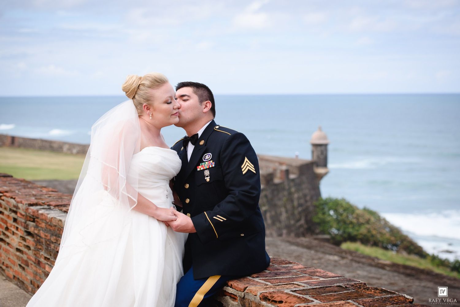 Military groom kisses bride in white wedding gown on historic brick fortress wall with ocean backdrop.