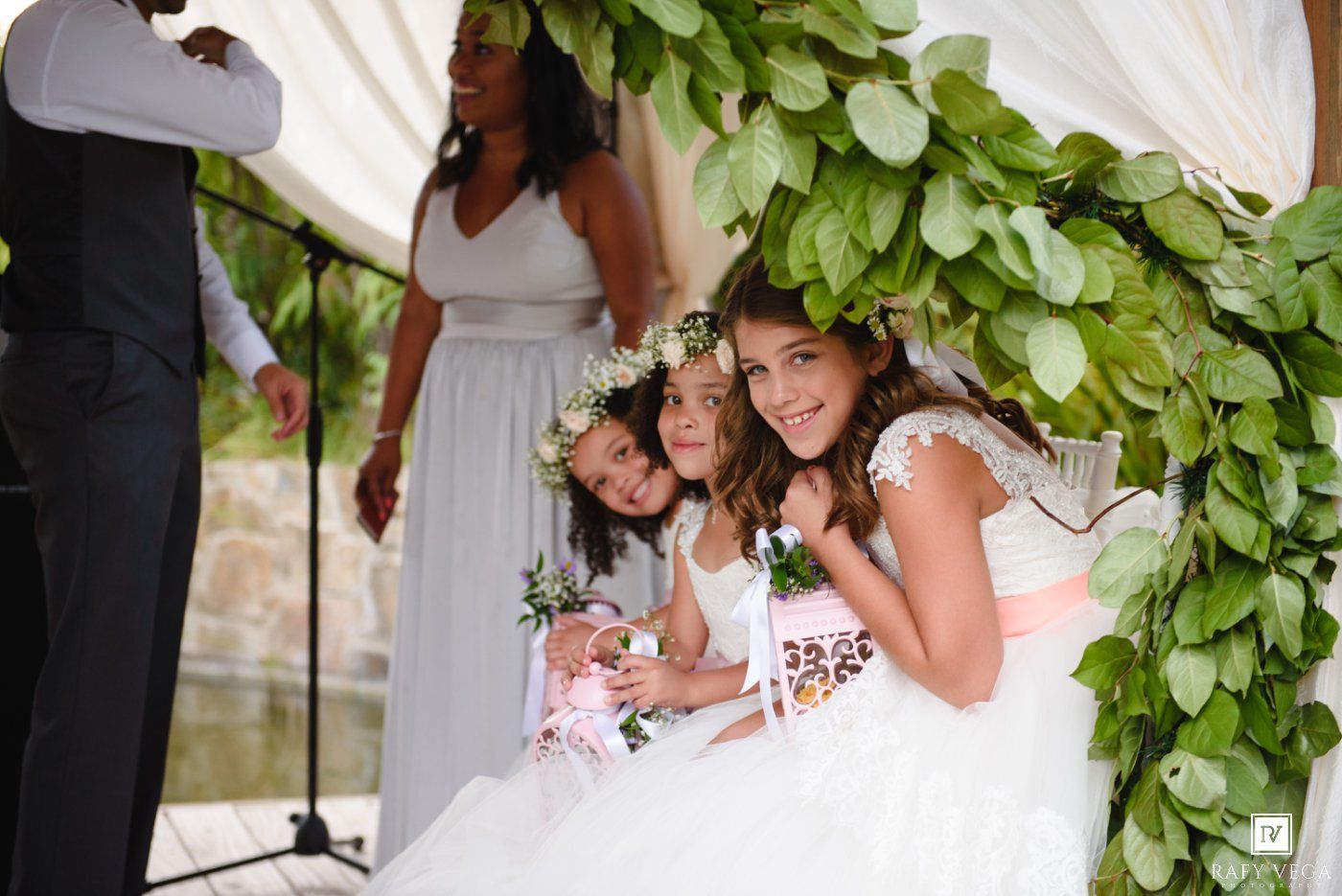Three young flower girls in white dresses and floral crowns pose together under a lush green leafy arch at an outdoor wedding.