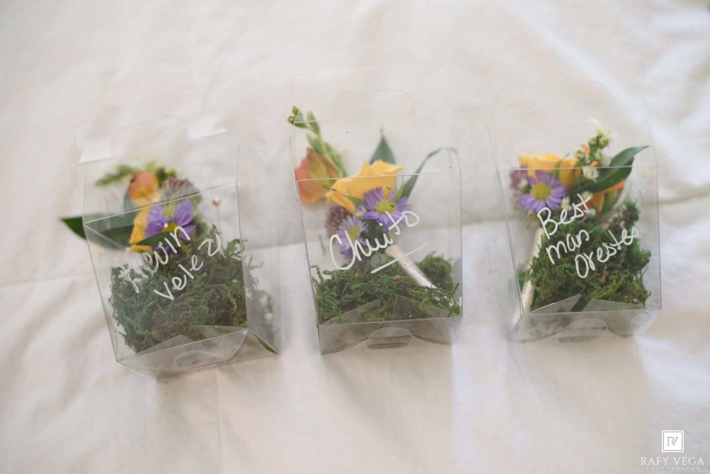 Three clear plastic bags filled with moss and colorful flowers with handwritten name tags on white fabric.