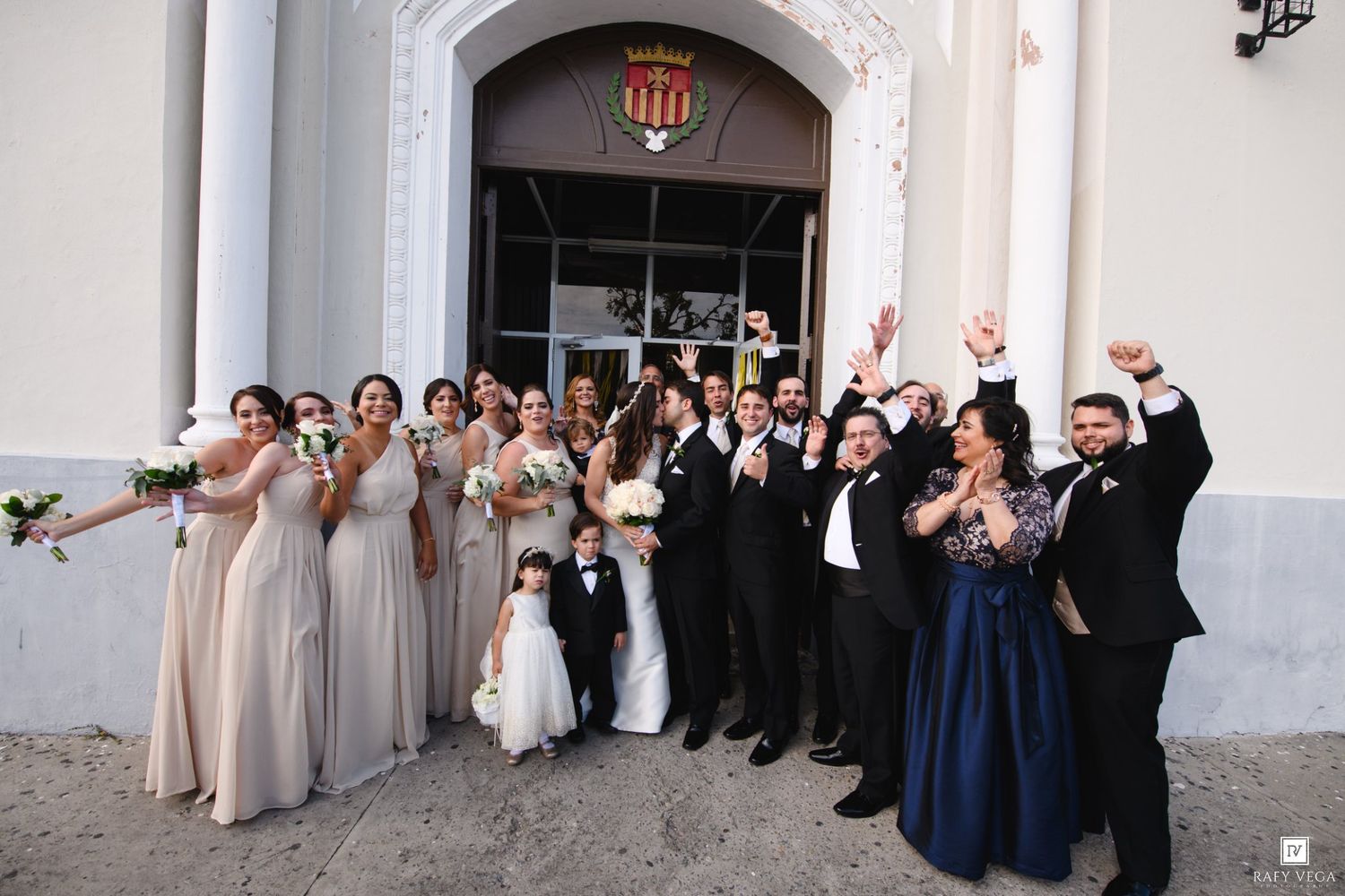 Wedding party celebrates outside church entrance with bridesmaids in champagne gowns and groomsmen in navy suits.