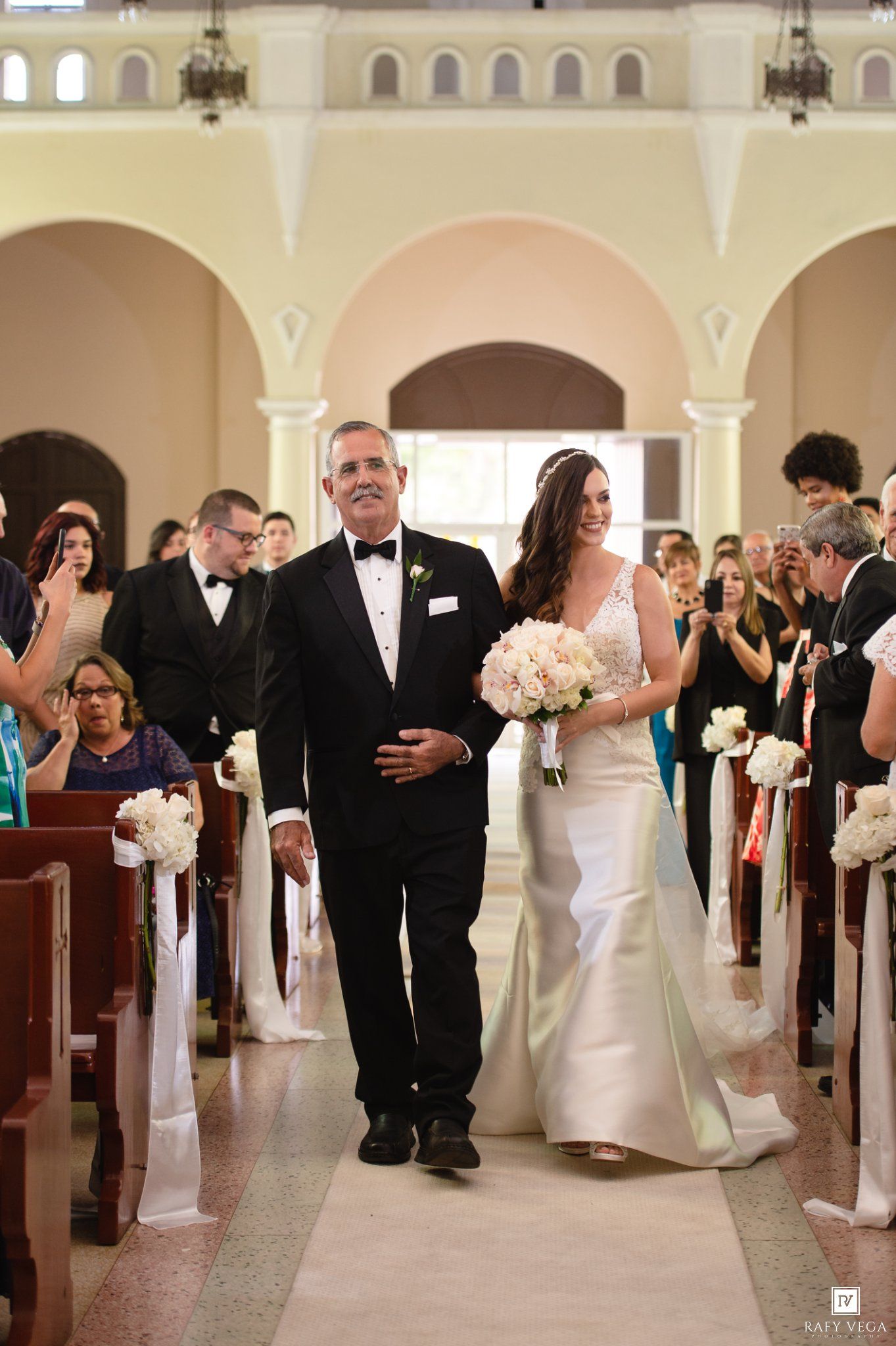 Bride walks down church aisle with father in tuxedo, holding white bouquet, guests standing in pews during wedding ceremony.