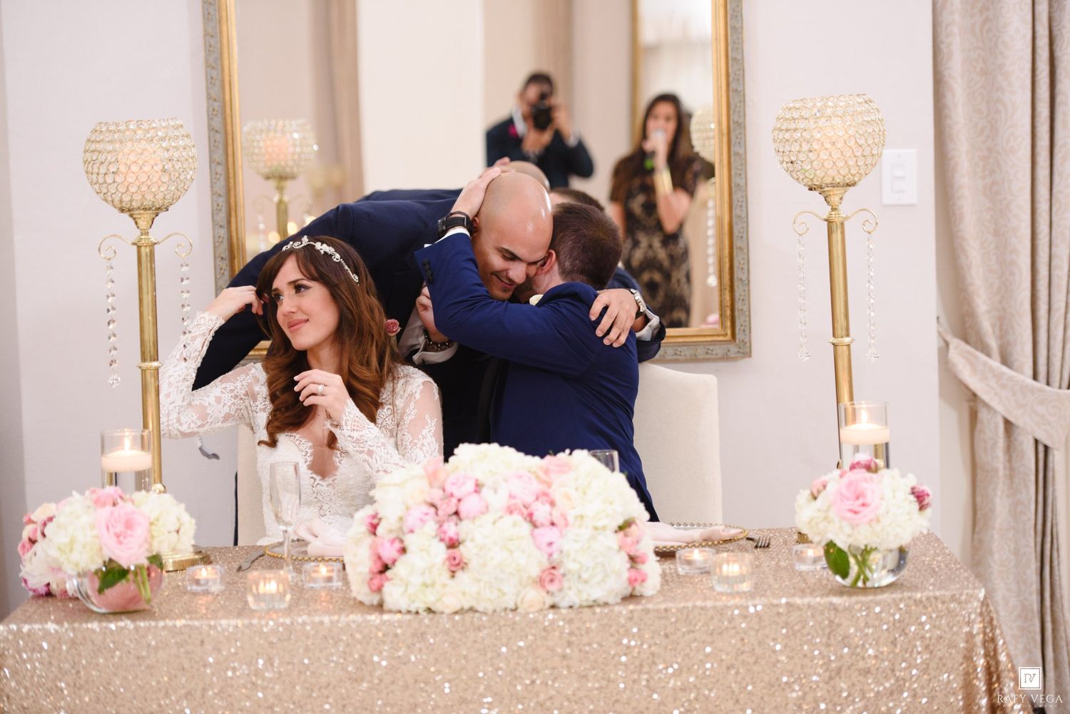 Emotional wedding reception moment with groom hugging guest at decorated head table with pink floral arrangements.