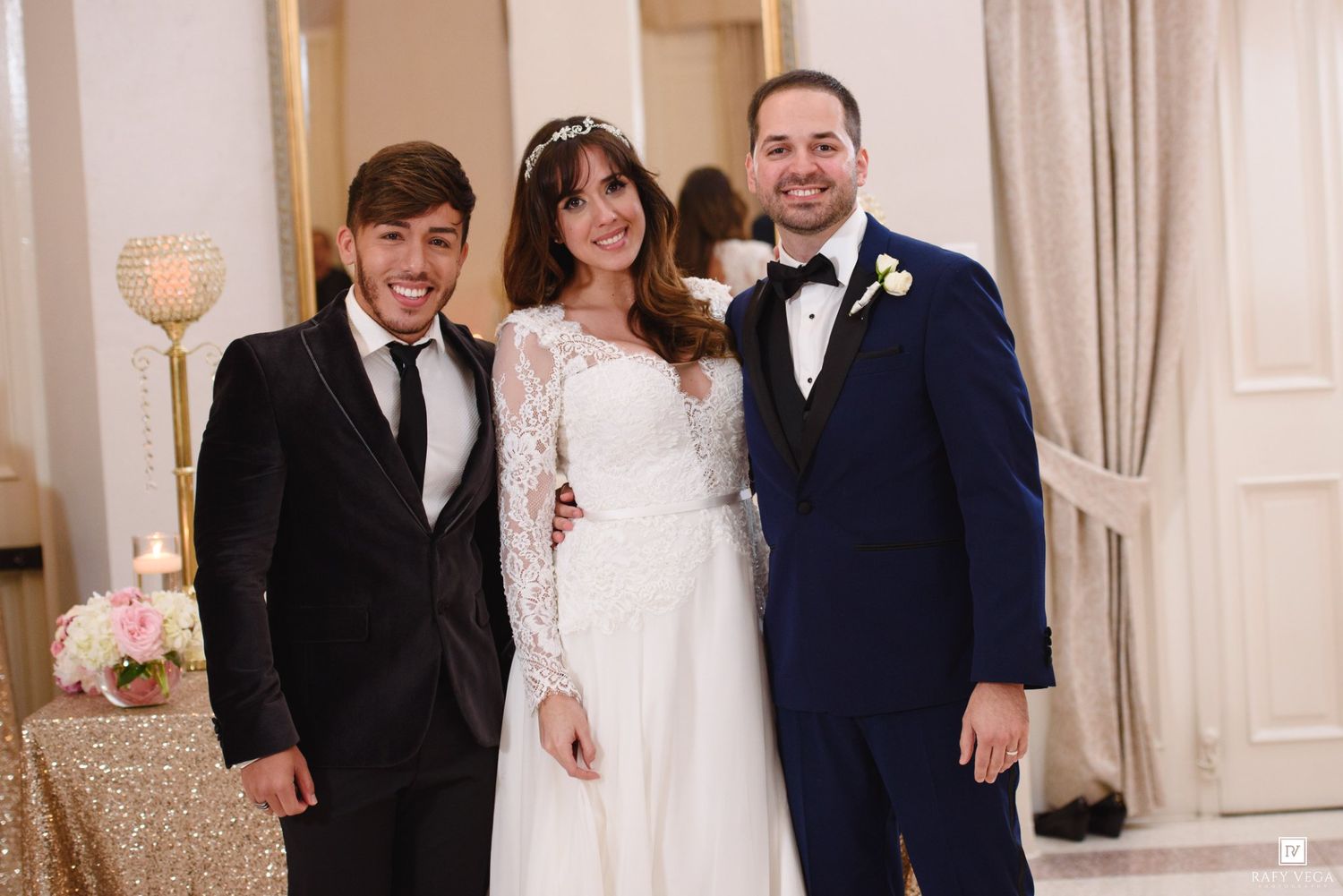 Bride in white lace gown poses with two men in suits at elegant wedding reception venue.