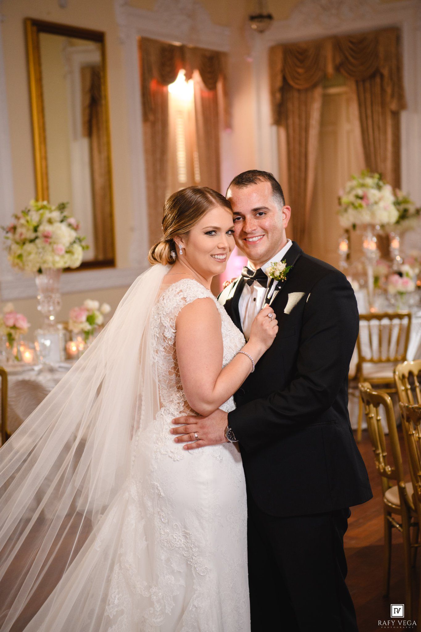 Bride in lace gown and groom in black tuxedo pose together at their elegant wedding reception venue.