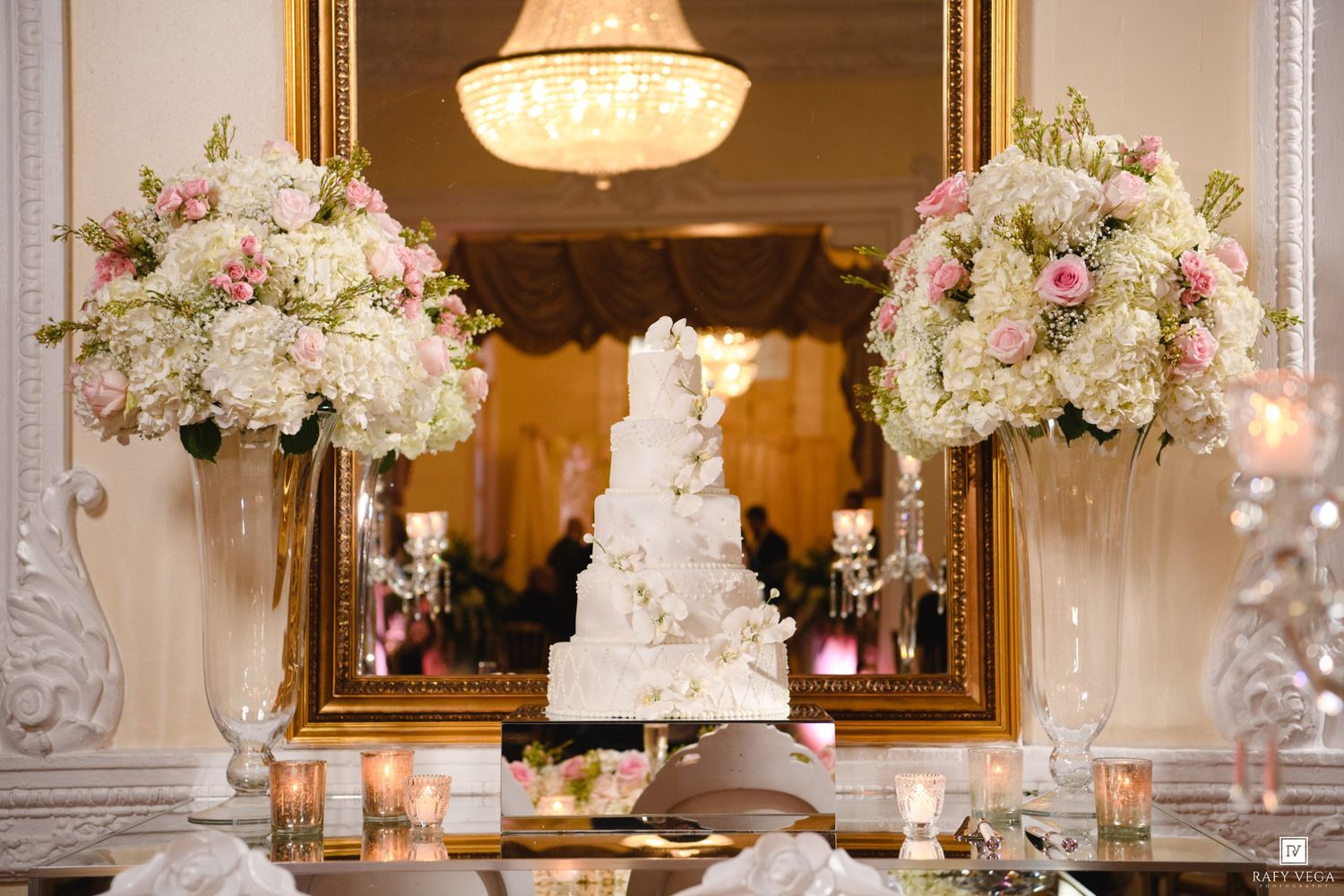 Elegant white tiered wedding cake displayed between tall floral arrangements of pink and white roses on a mirrored table.