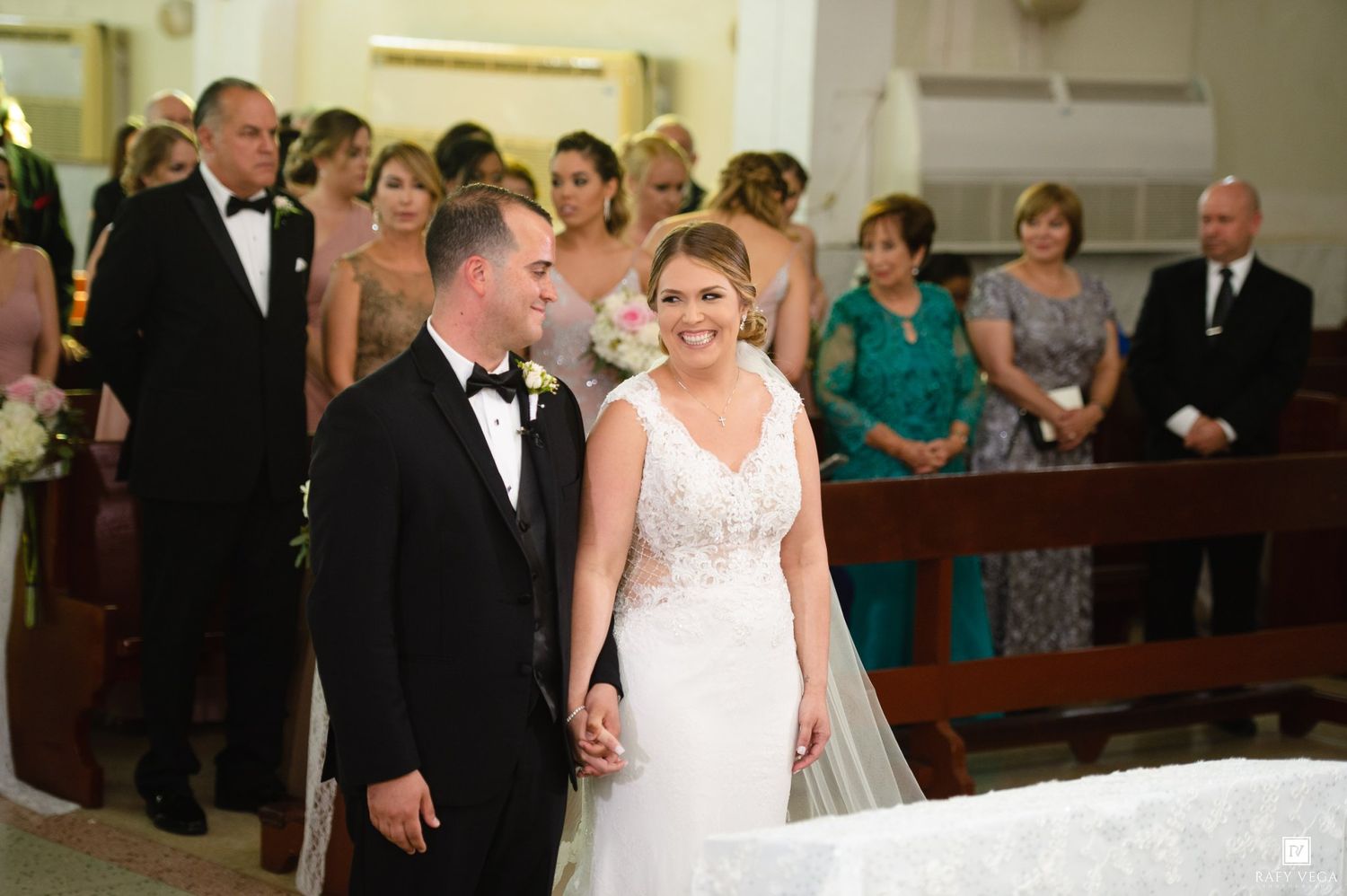 Bride and groom holding hands at church altar, smiling, with bridesmaids and guests in background.