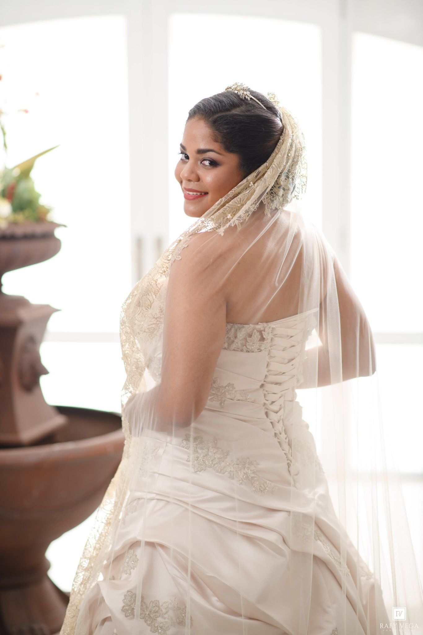 Smiling bride in strapless lace wedding gown and veil poses elegantly near a fountain in a bright venue.