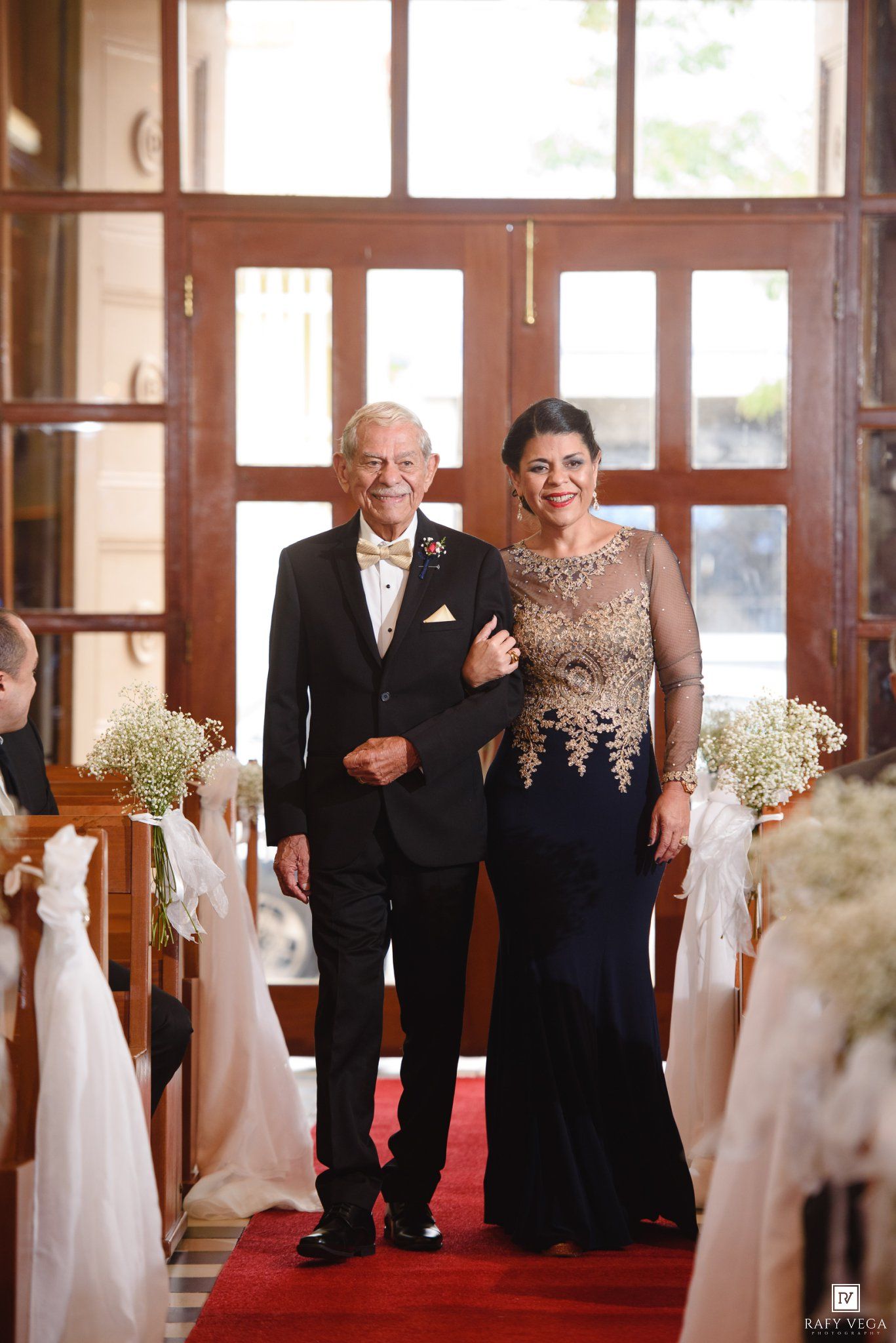 Elegant couple walking down wedding aisle, man in tuxedo and woman in navy gold gown, church ceremony with floral decorations.