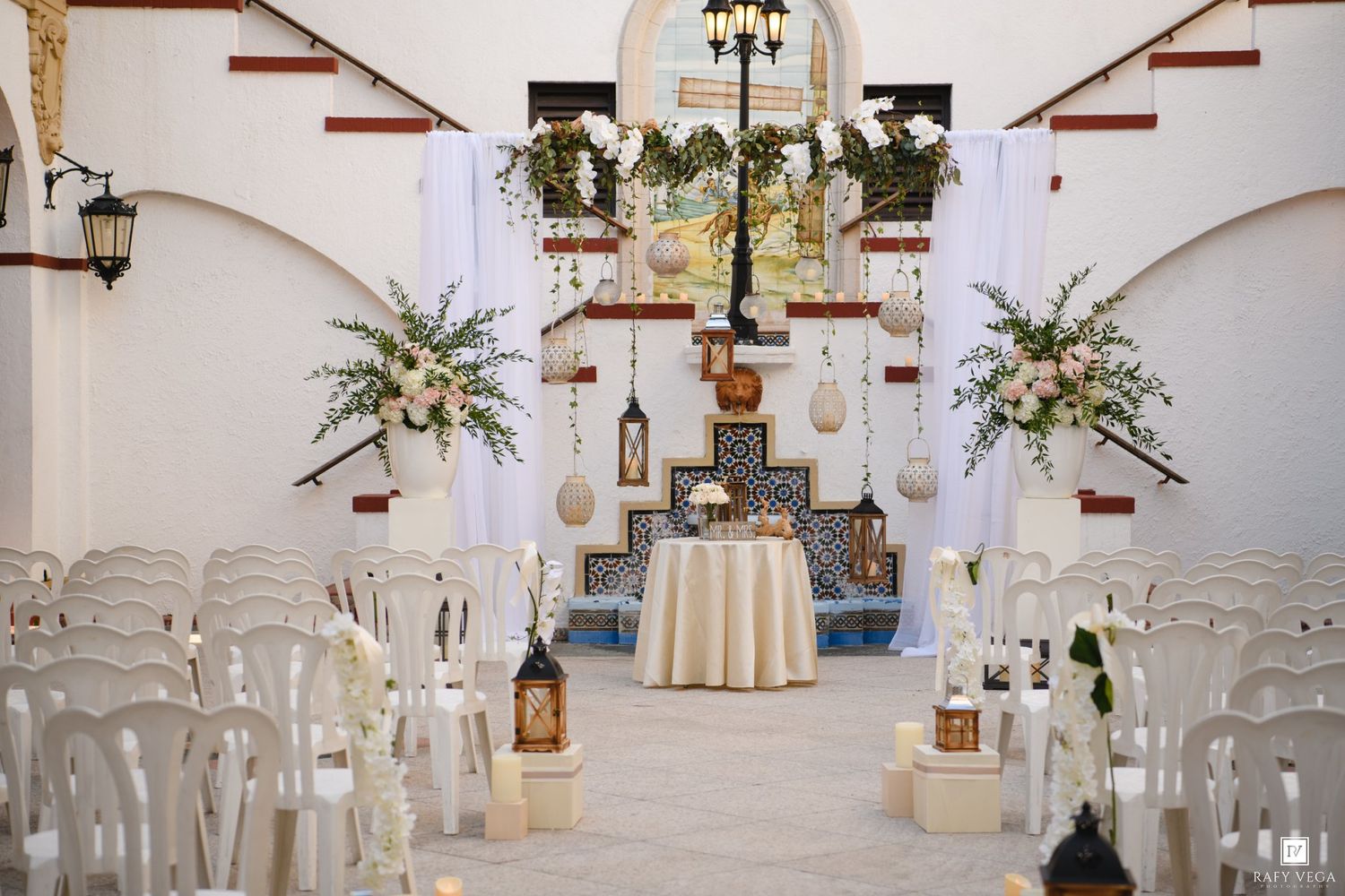 Elegant wedding ceremony setup with white chairs, floral arrangements, lanterns, and a decorated altar in a white venue.