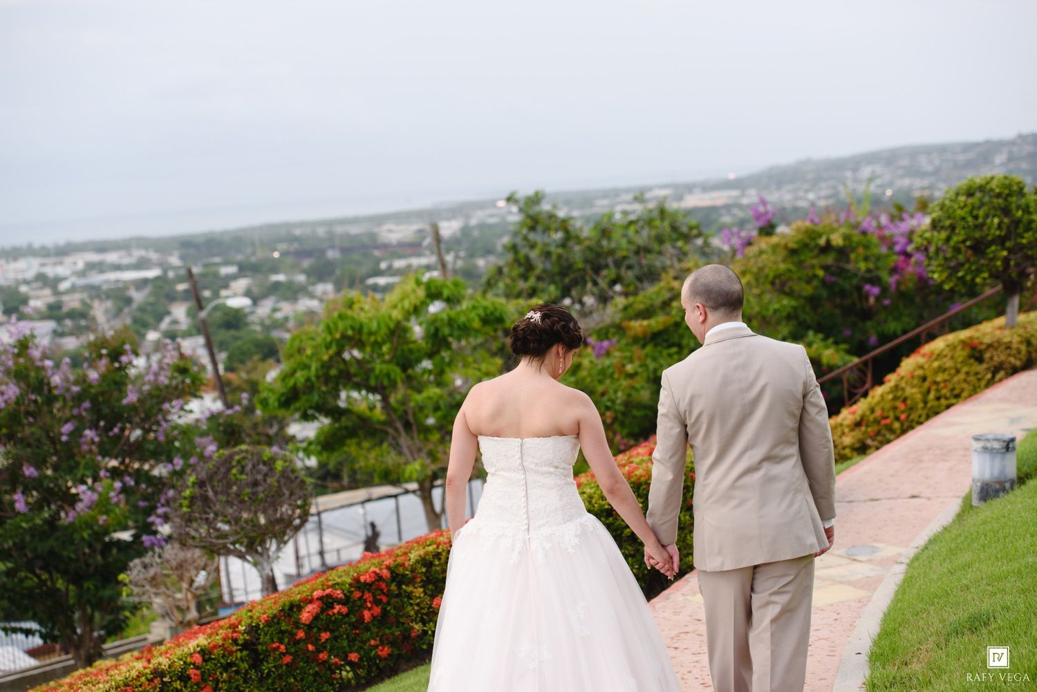 Bride and groom hold hands walking along a garden path with a scenic coastal city view in the background.