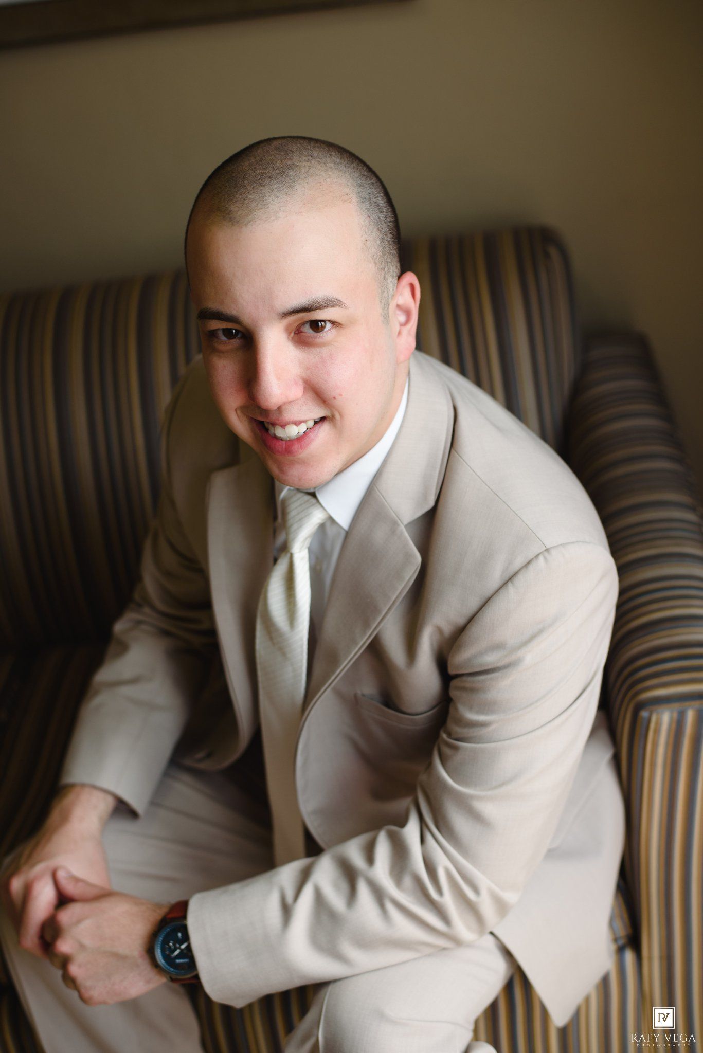 A smiling young man in a beige suit and tie sits relaxed in a striped armchair, posing for a formal portrait photo.