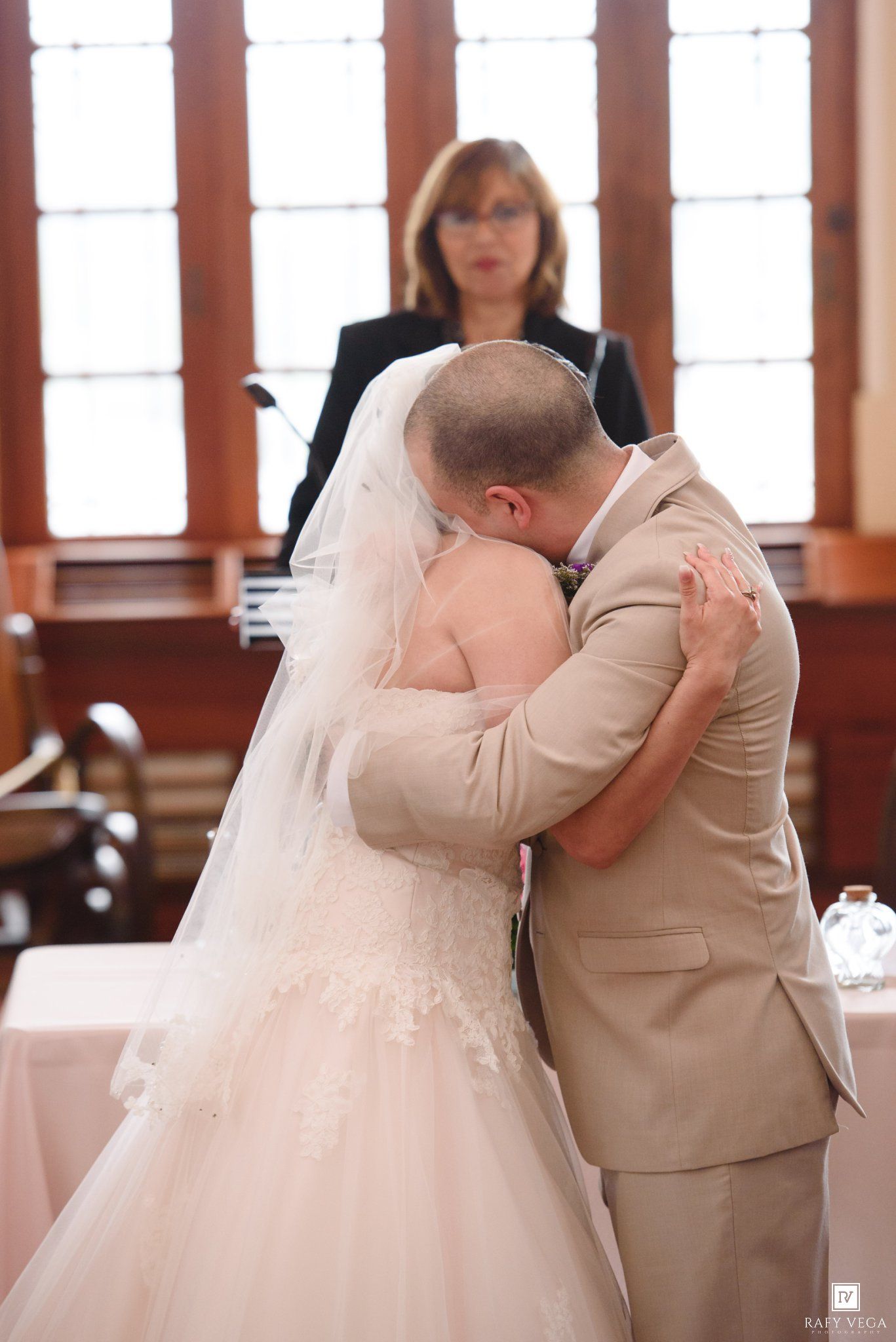 Bride and groom share first kiss at wedding ceremony while officiant watches in elegant venue.
