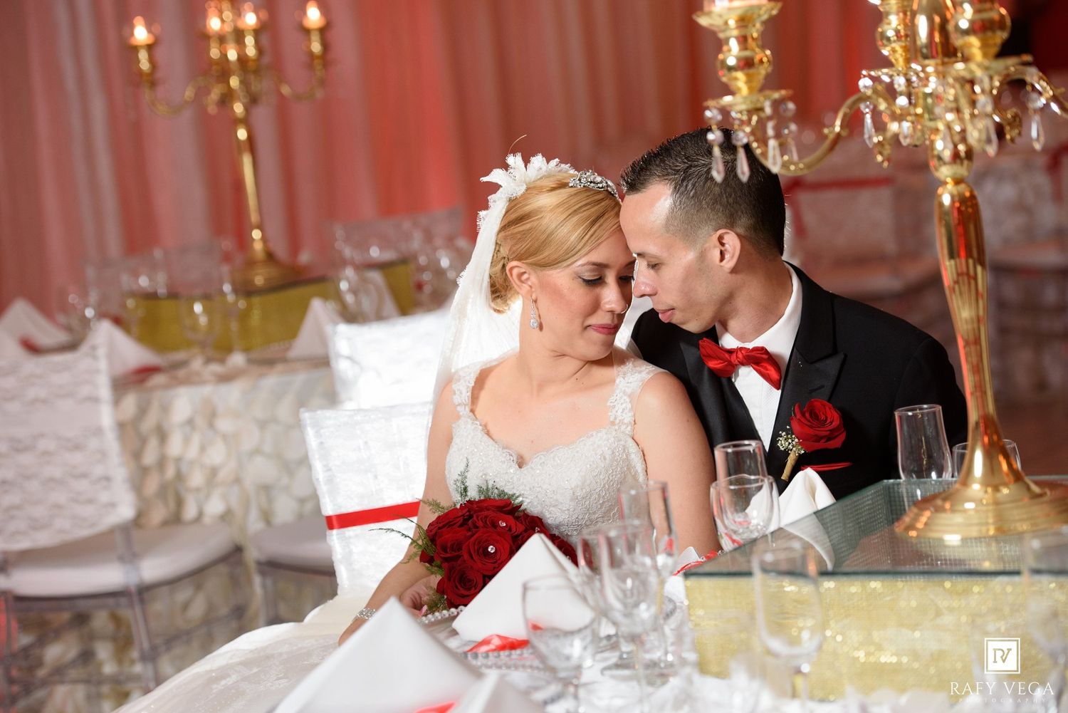 Bride and groom share an intimate moment at their wedding reception, surrounded by red roses and elegant gold candelabras.