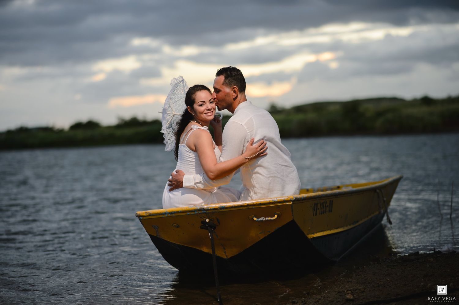 A bride and groom share a romantic embrace in a yellow boat on a calm lake under dramatic cloudy skies.