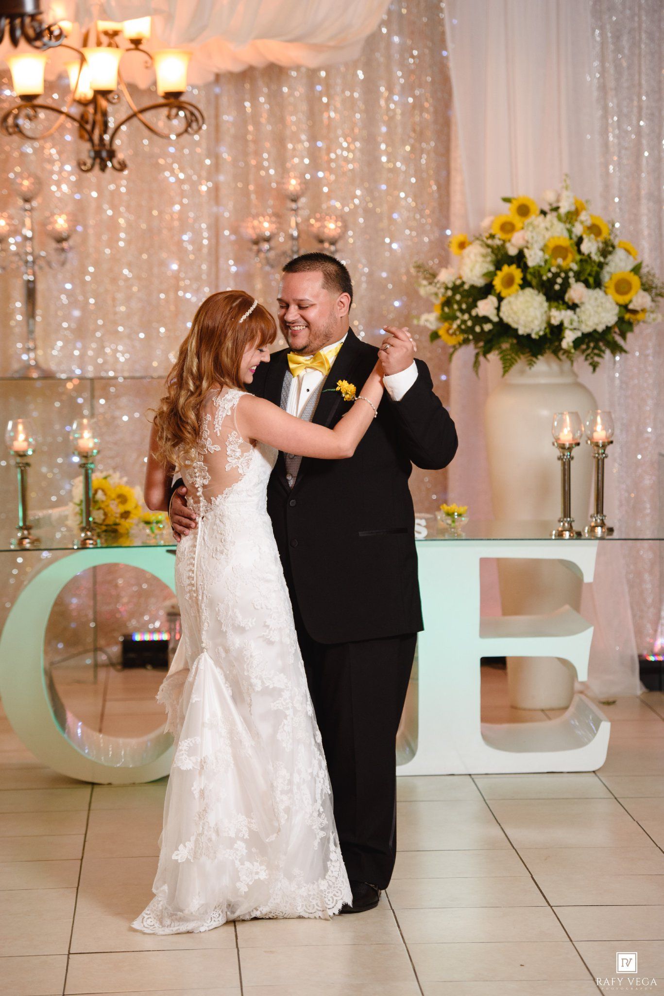Bride and groom sharing first dance at elegant wedding reception with sparkle backdrop and floral arrangements.