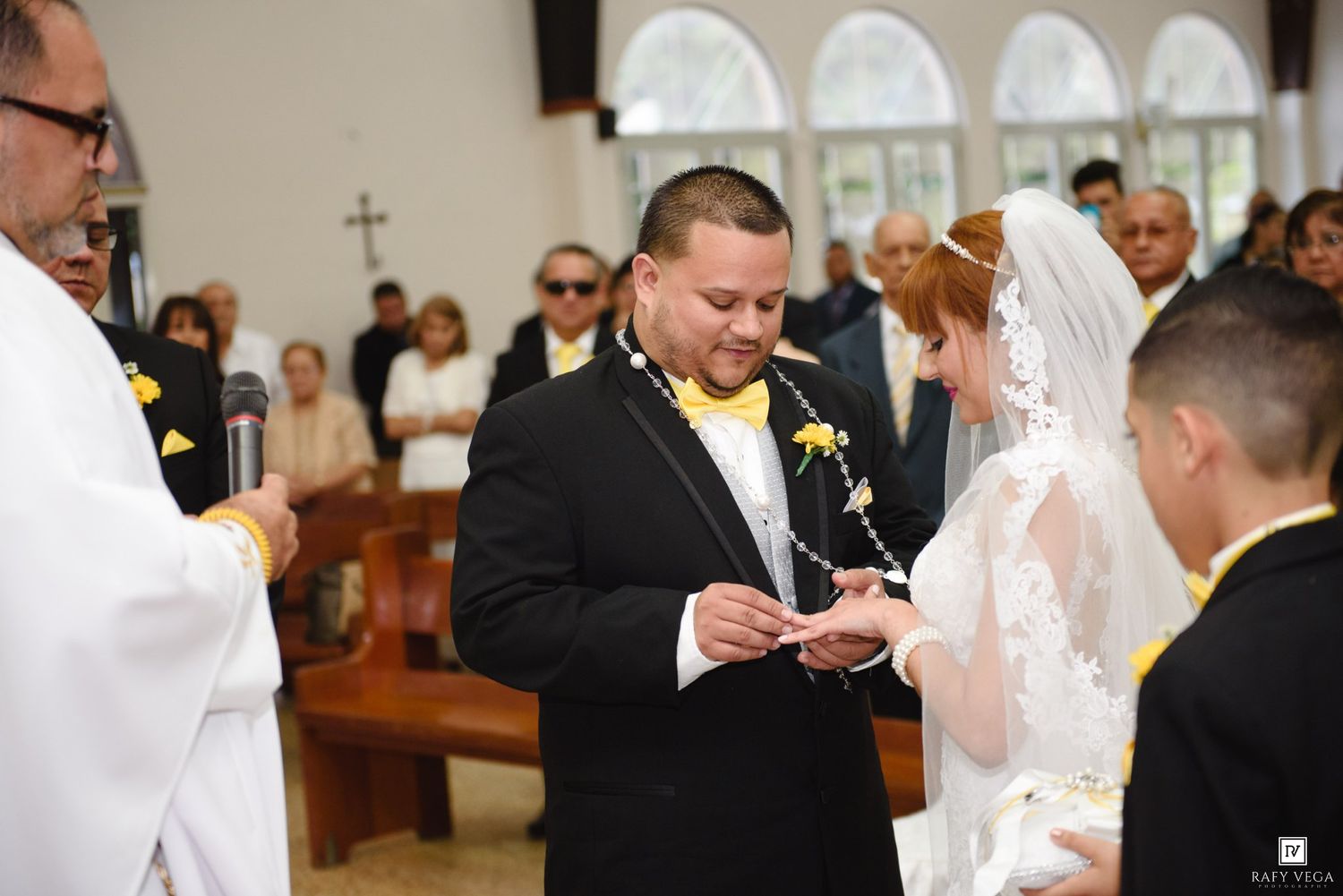Groom in black tuxedo placing wedding ring on bride in white veil during church ceremony with yellow floral accents.