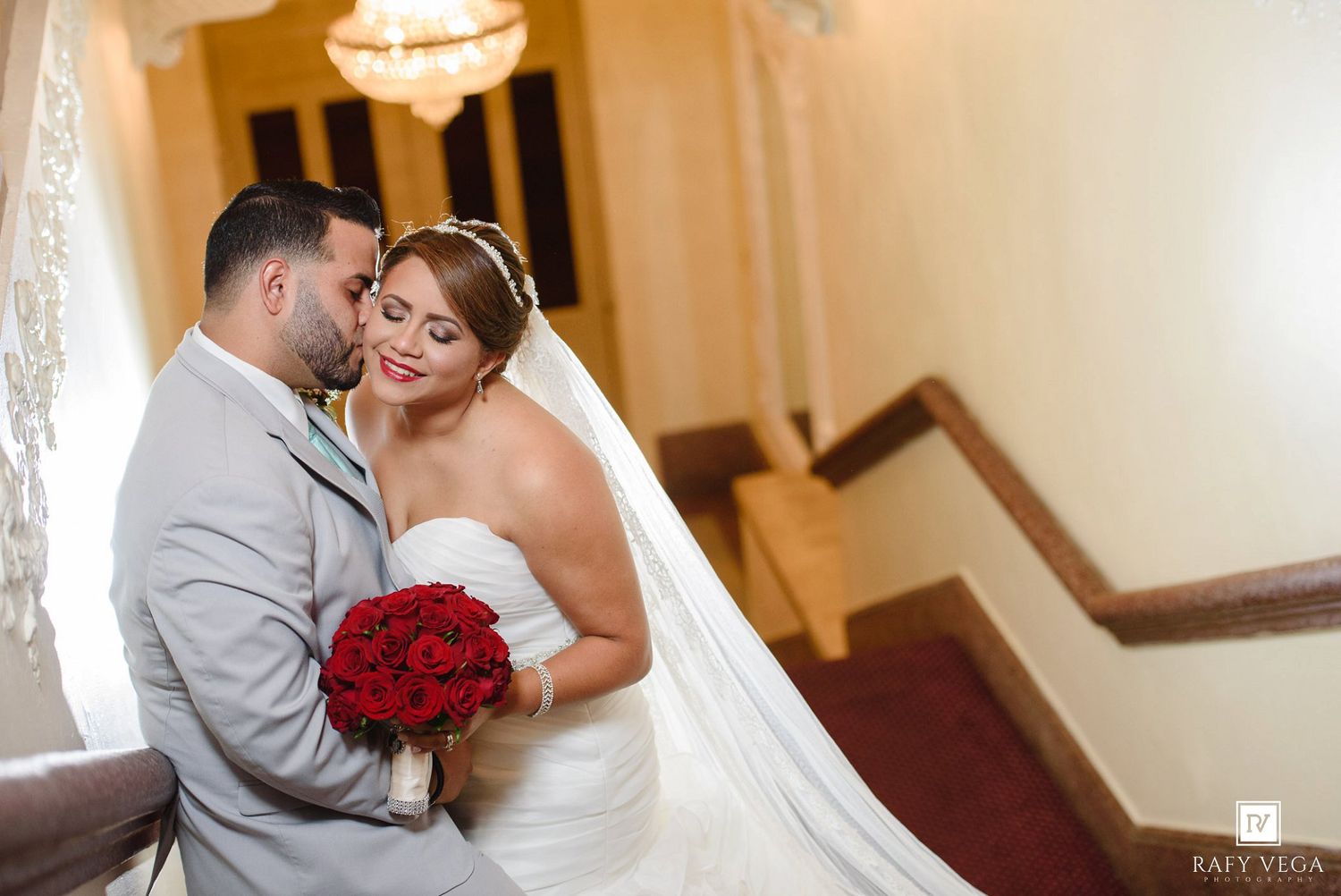 Bride and groom share a romantic embrace on a staircase, bride holding a red rose bouquet in a white gown.