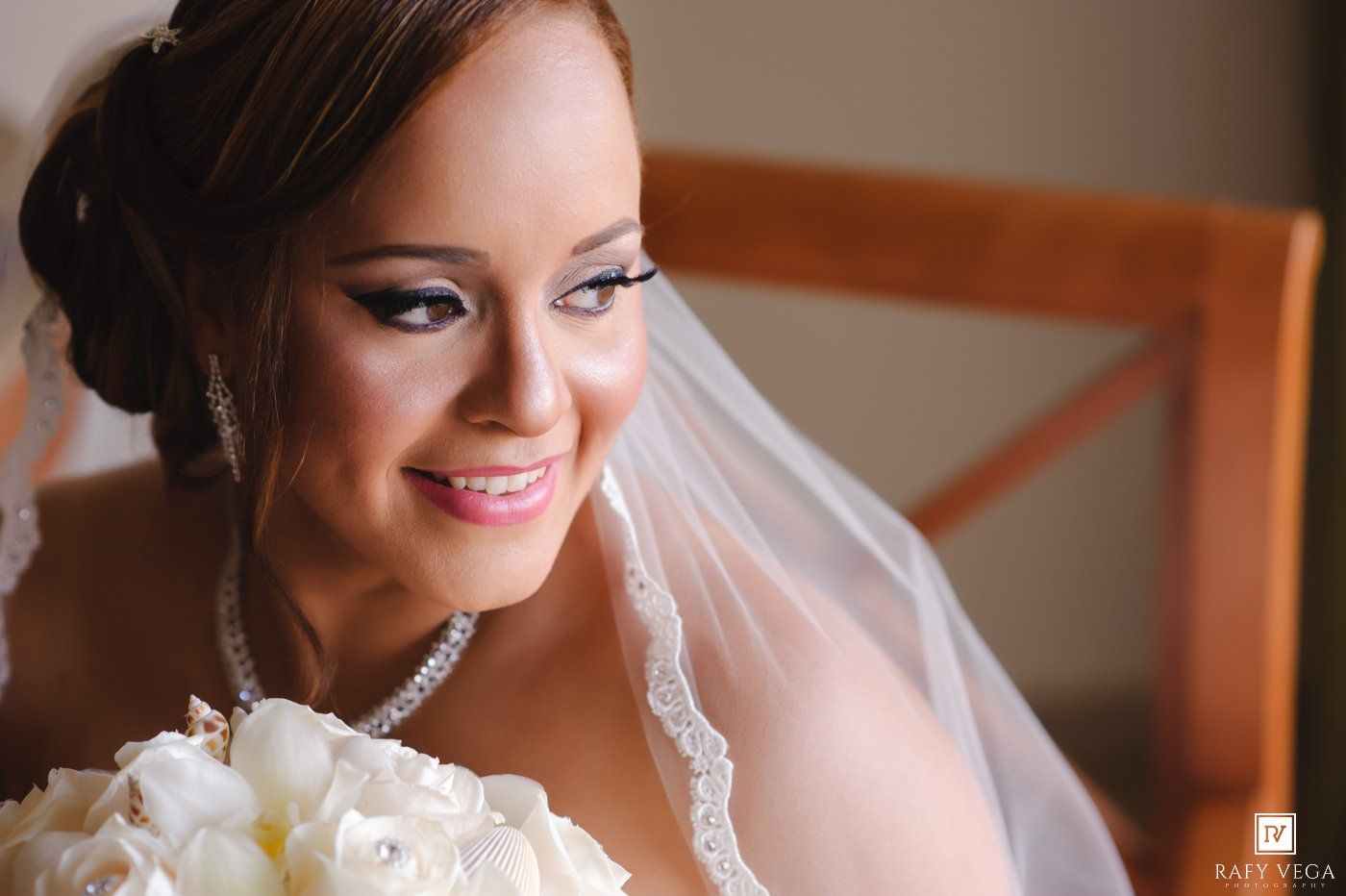 Smiling bride in wedding veil holding white bouquet with elegant updo hairstyle and pearl necklace on her wedding day.