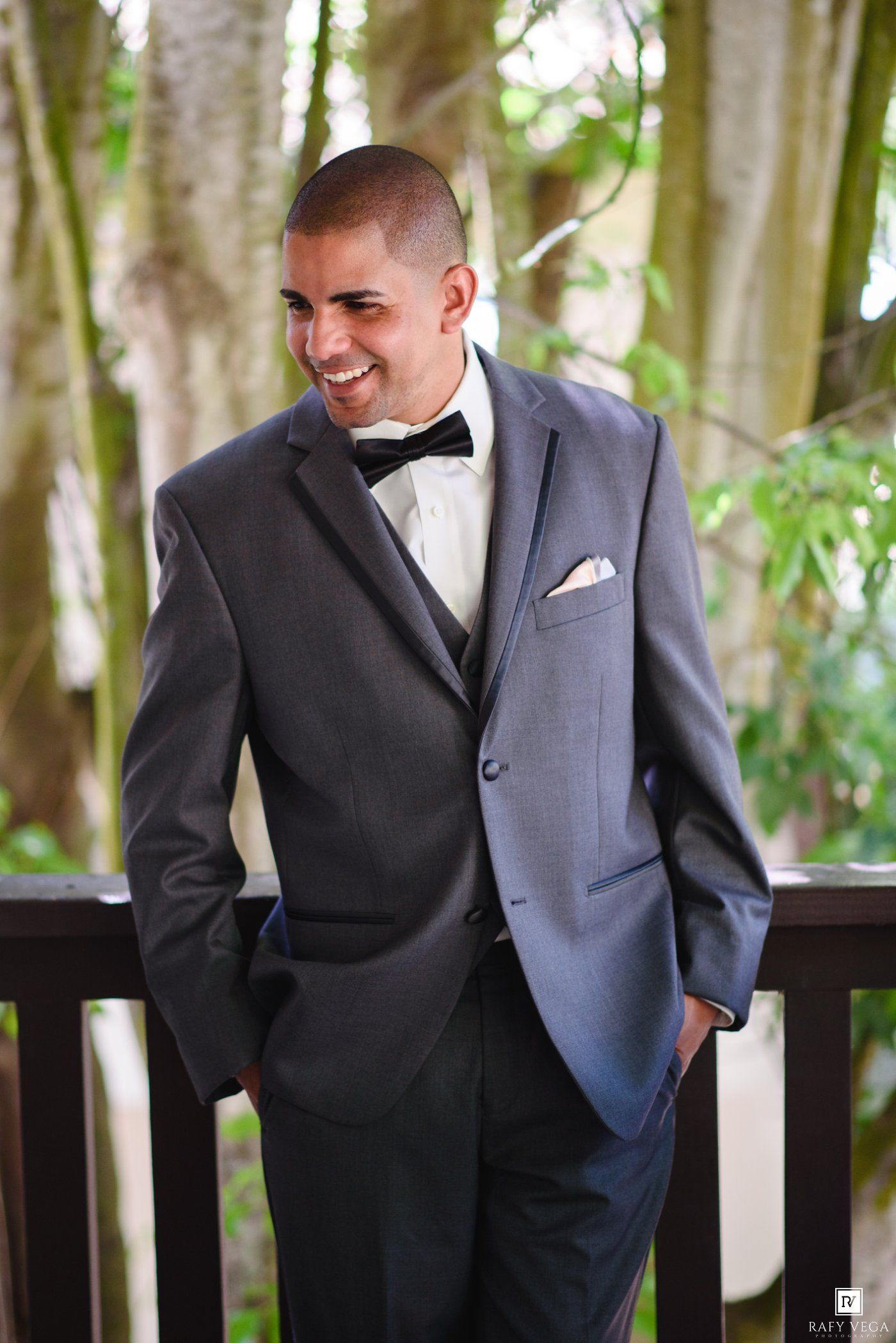 Smiling groom in gray tuxedo with black bow tie and pocket square stands on wooden deck surrounded by trees.