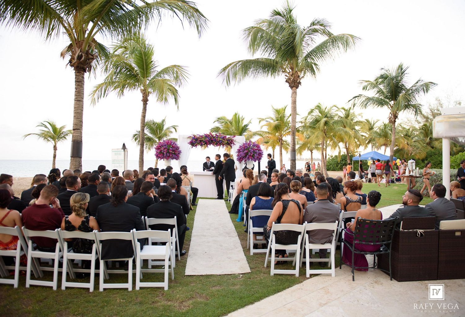 Outdoor beach wedding ceremony with guests seated in white chairs under palm trees with purple floral arrangements.