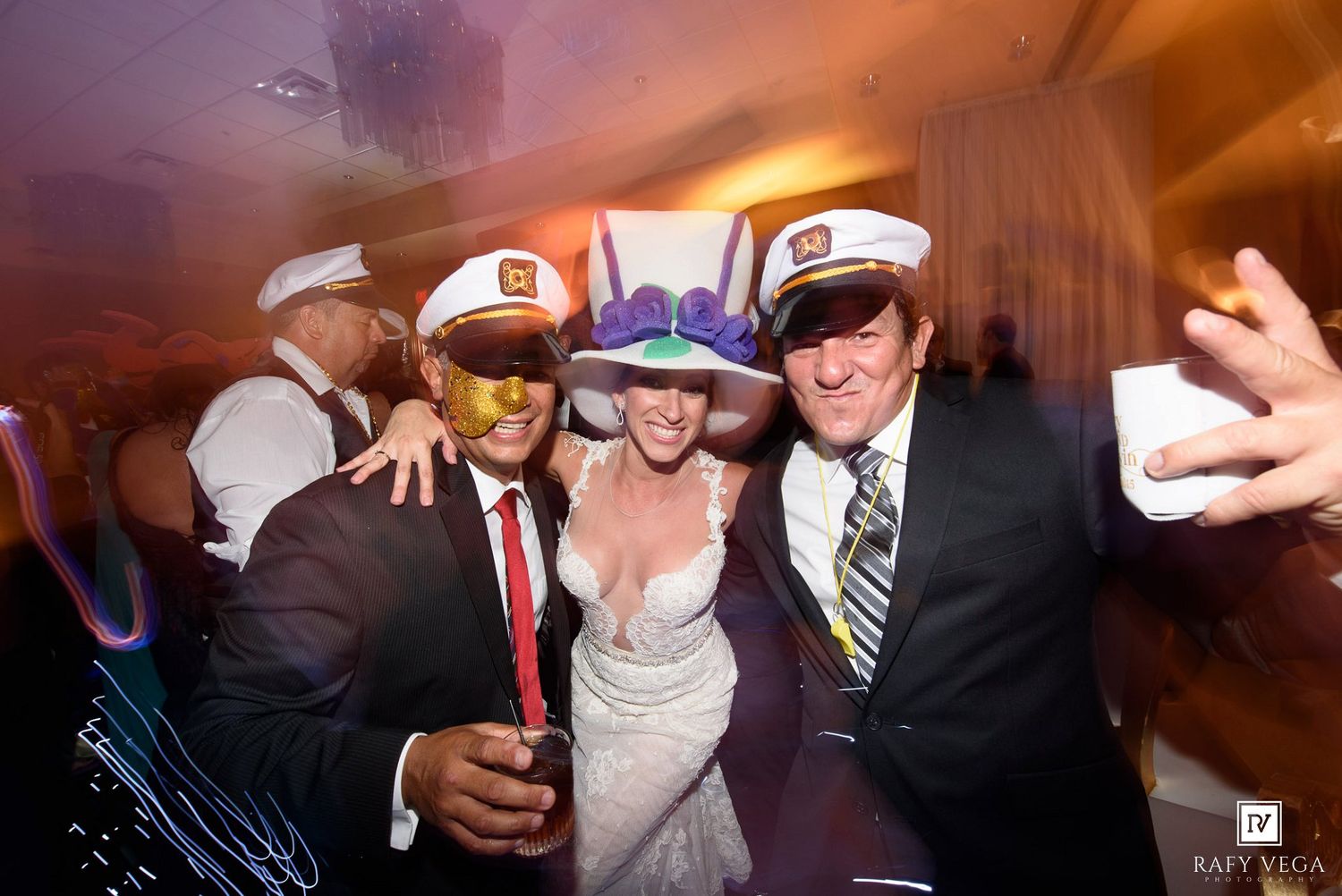 Three party guests wearing captain hats smile and pose together at a festive wedding reception celebration.