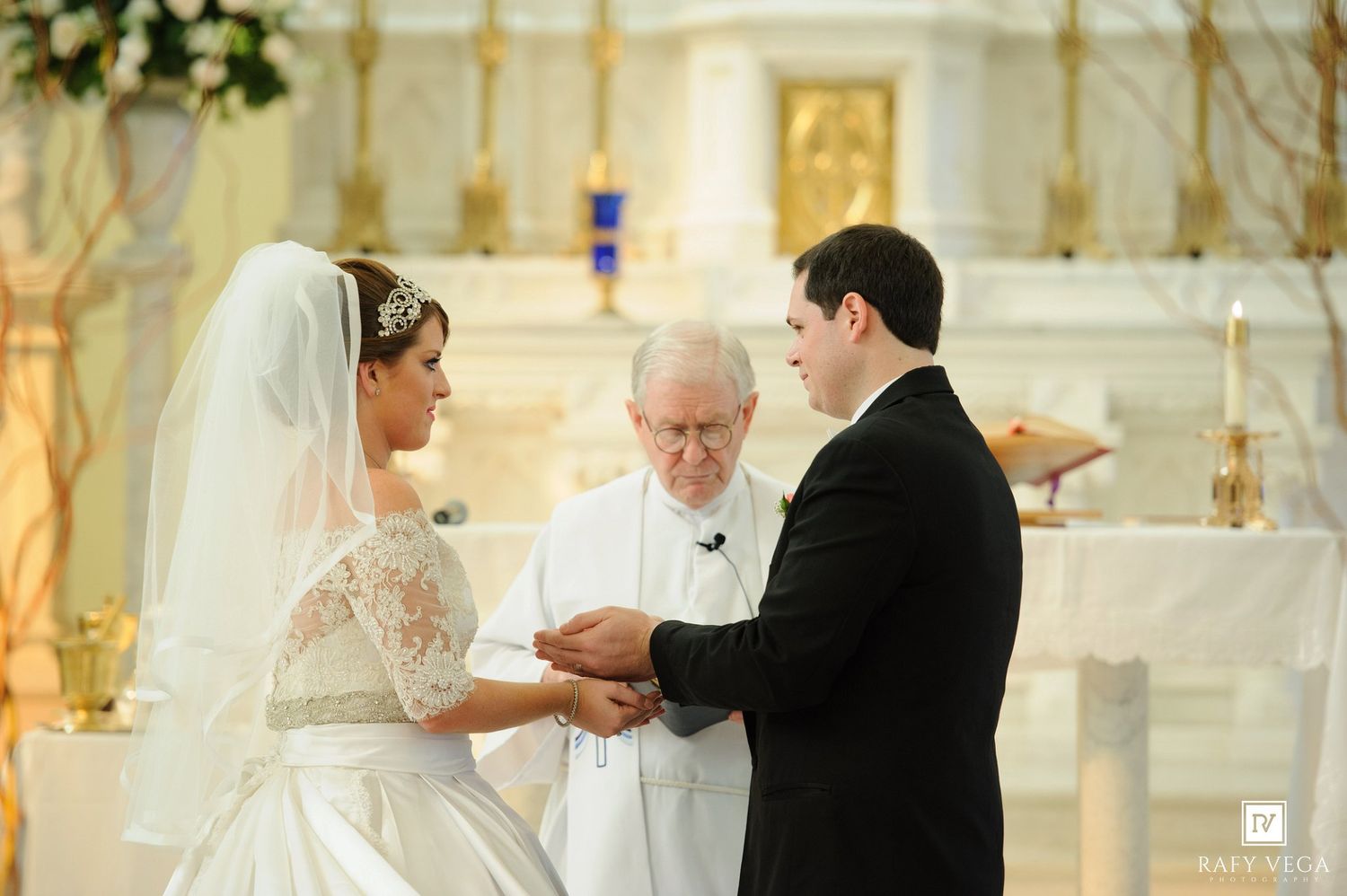 Bride and groom exchange rings at church altar with officiant during elegant Catholic wedding ceremony.