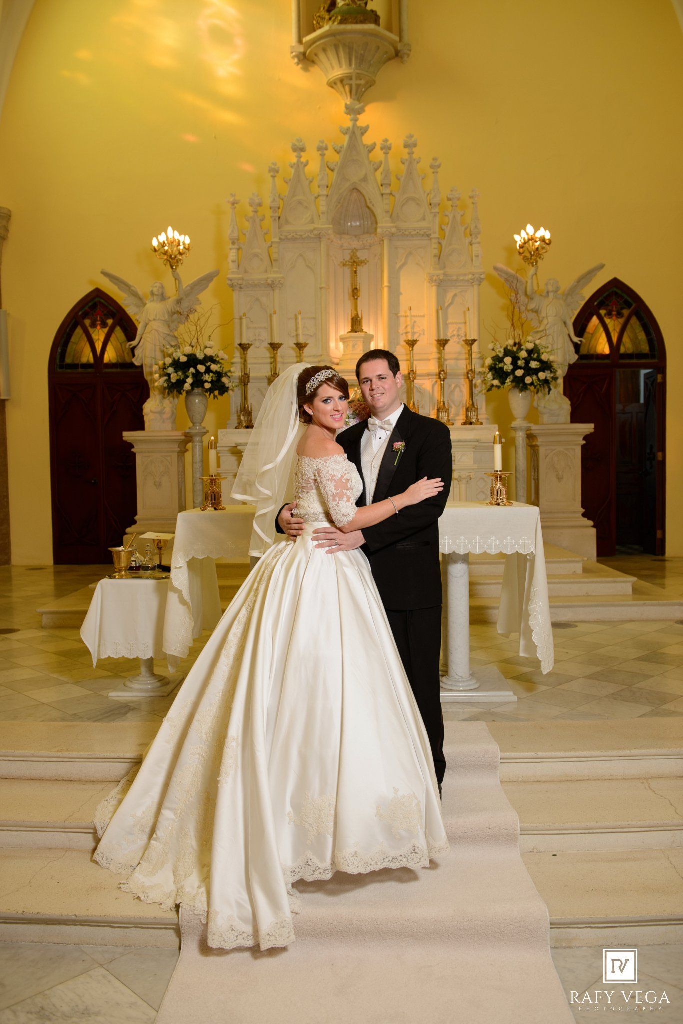 Stunning bride in ivory ball gown and groom in black tuxedo posing before ornate white church altar on wedding day.