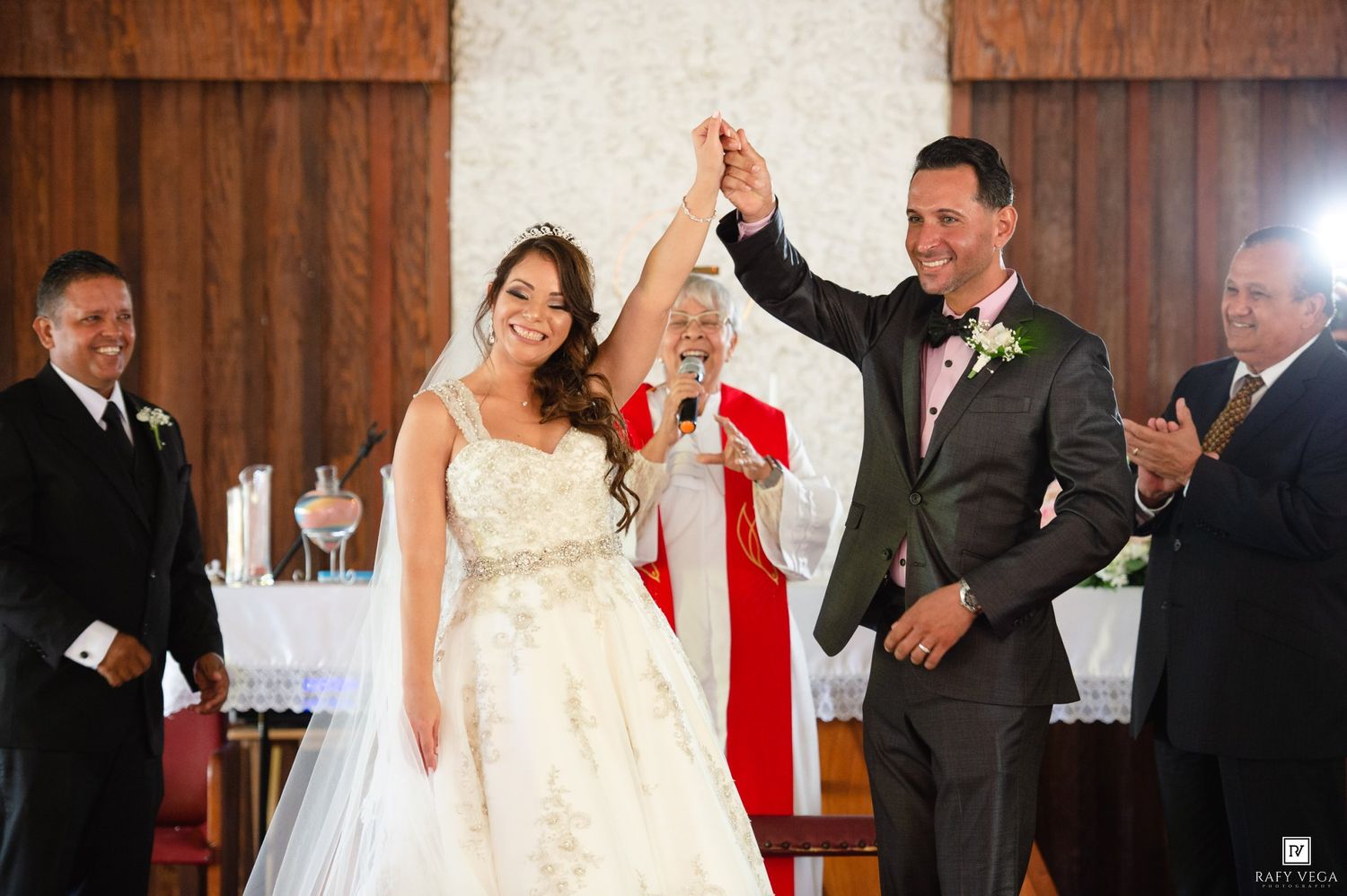 Joyful bride and groom raising hands at church wedding ceremony altar with officiant in red robes.