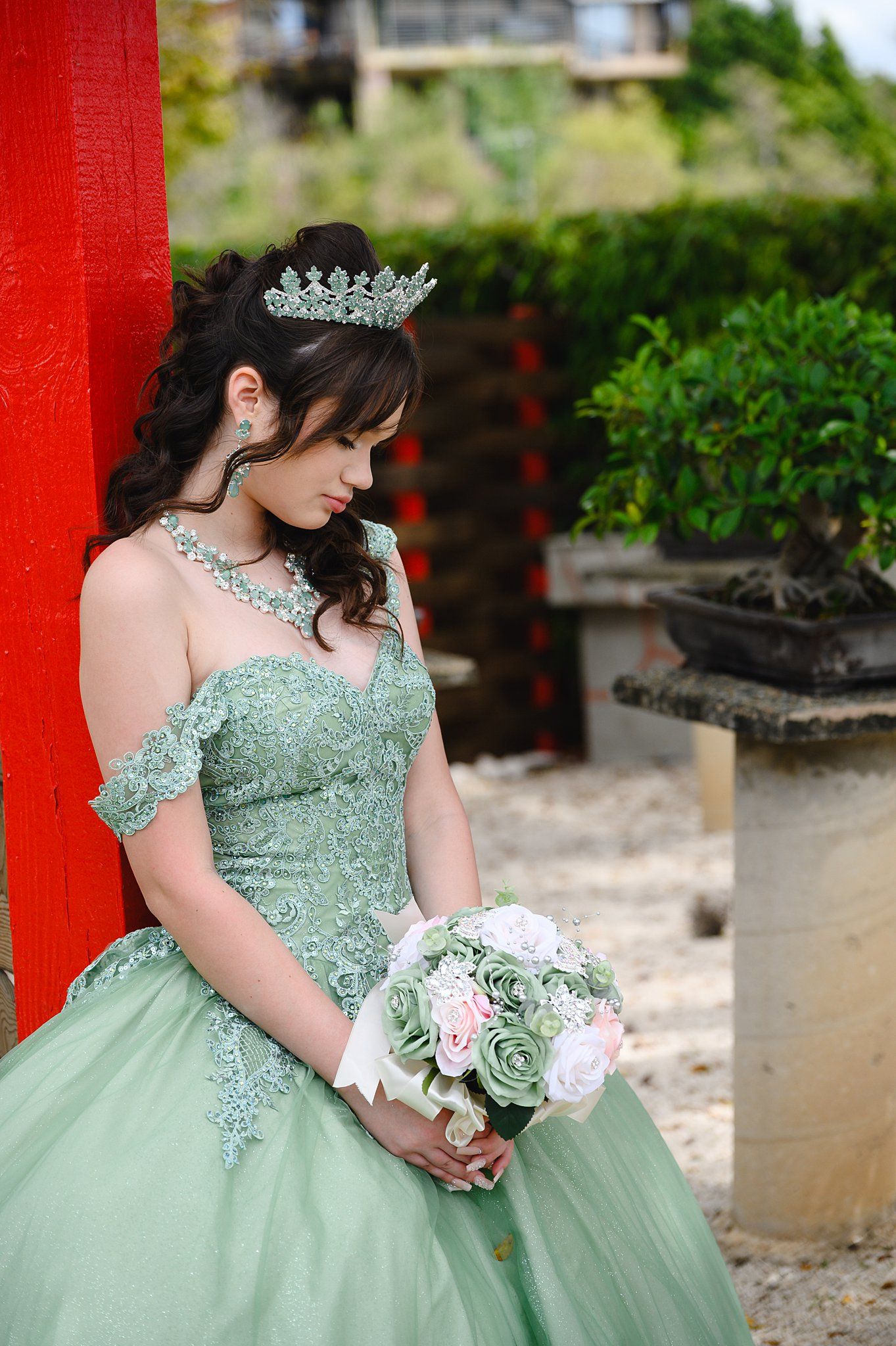 Young woman in sage green ball gown with tiara holds floral bouquet, posing elegantly in a garden setting.