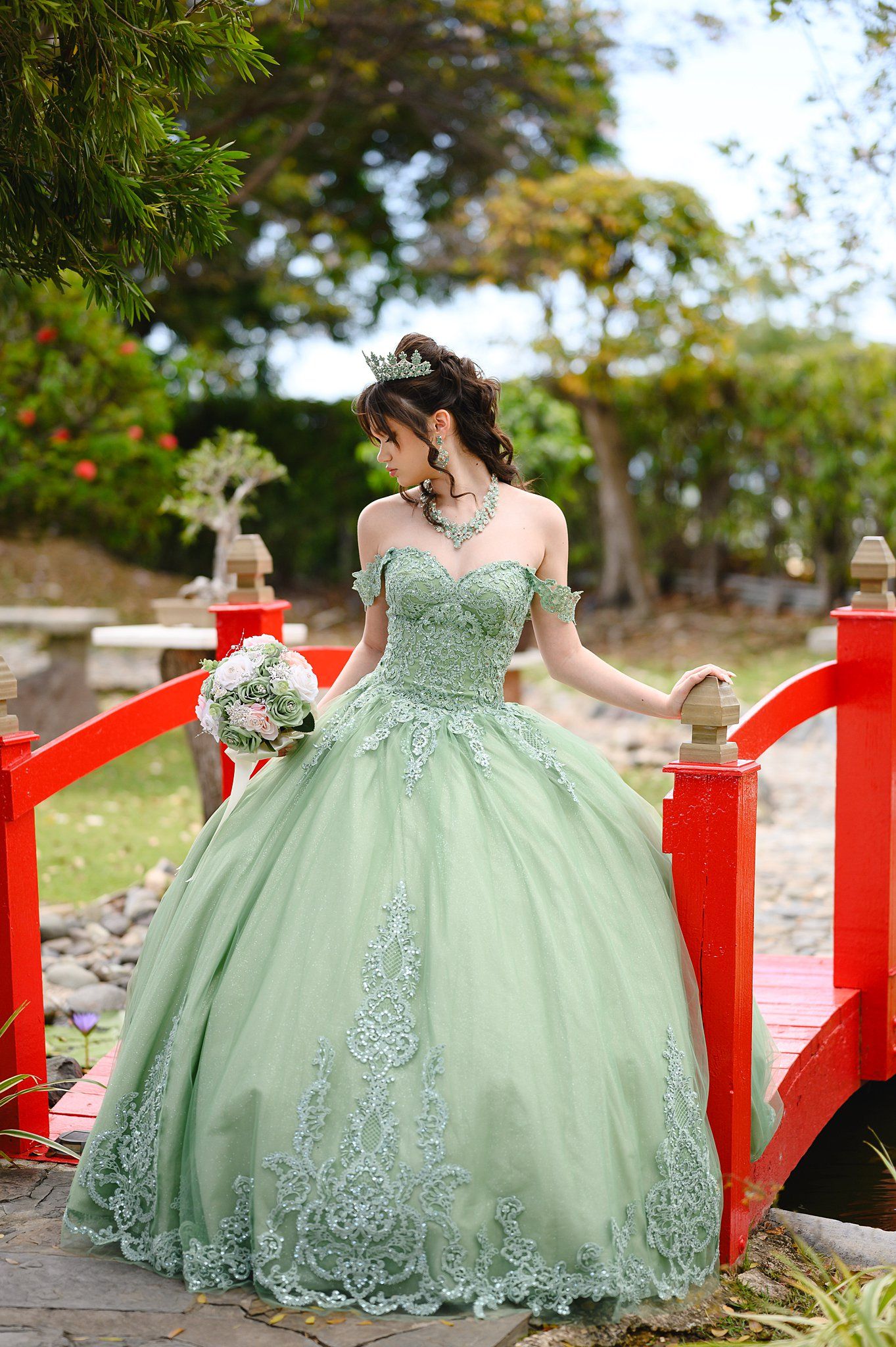 Young woman in sage green off-shoulder ball gown with lace appliques poses on red bridge holding bouquet.