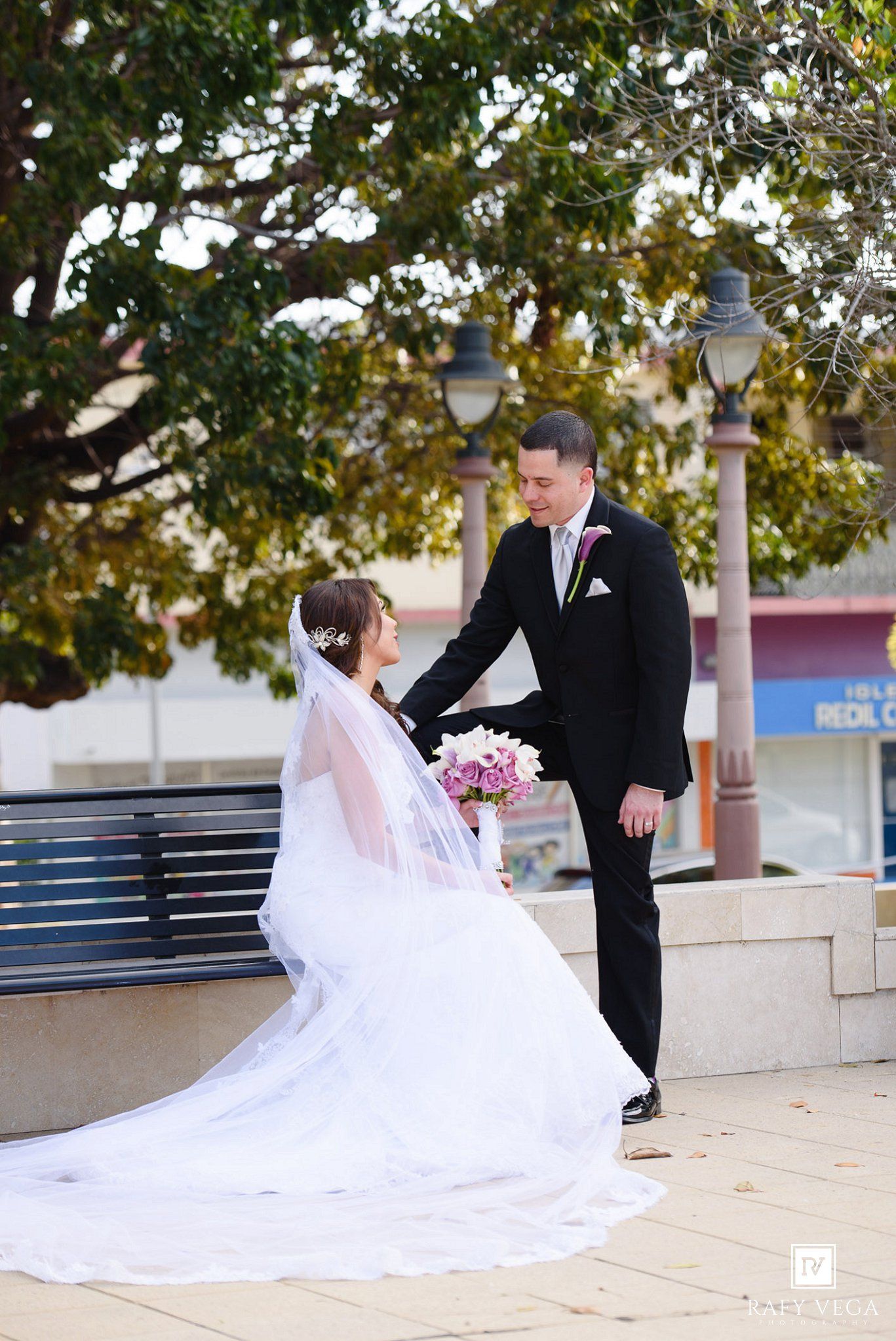 Bride in white gown sits on bench holding pink bouquet as groom in black suit reaches for her hand outdoors.