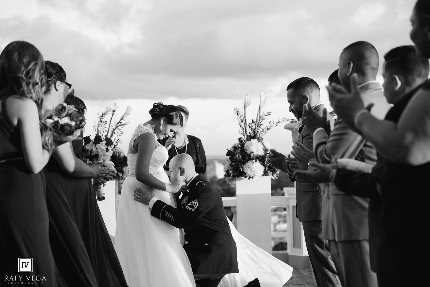 Black and white photo of bride and groom sharing first kiss at outdoor wedding ceremony with wedding party.